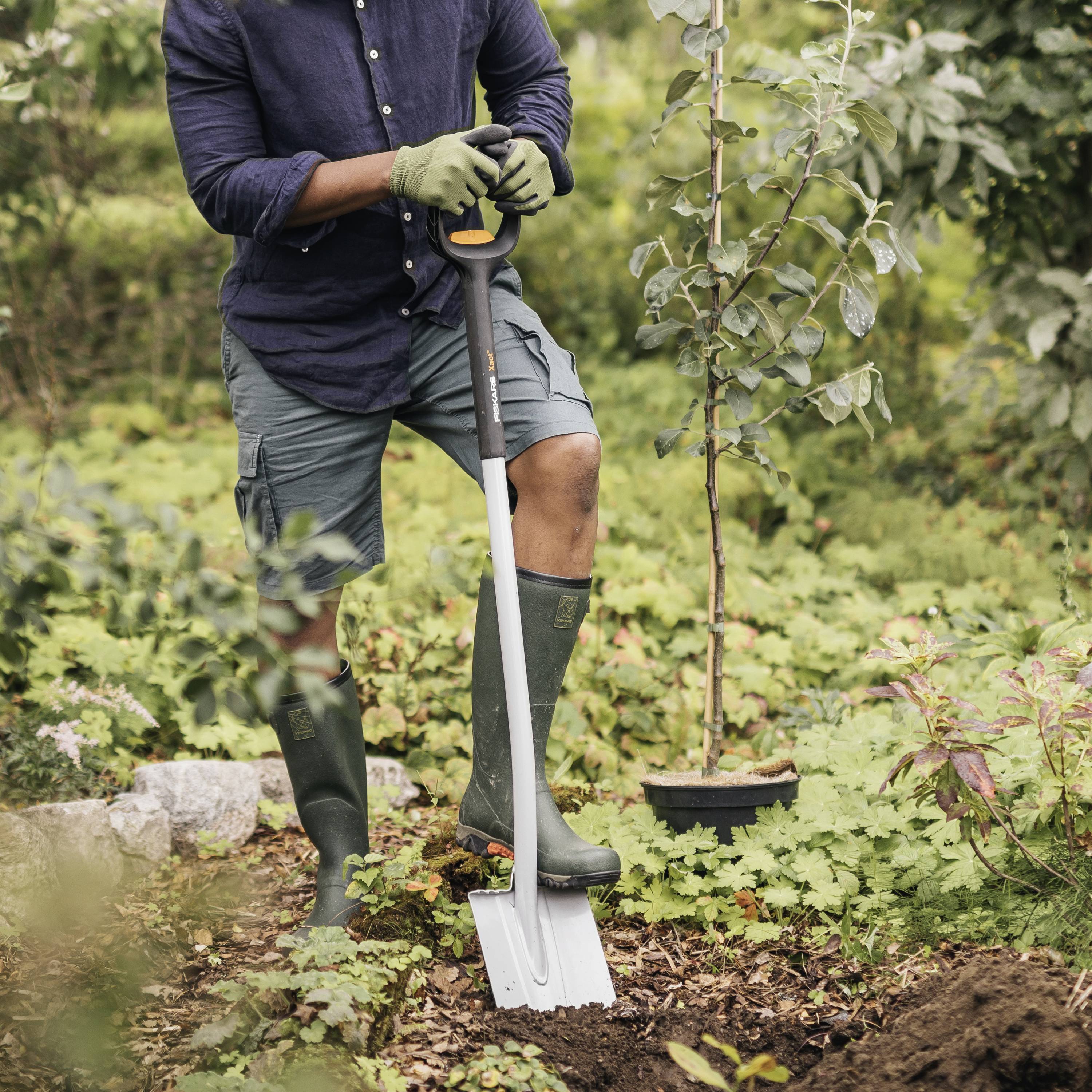 Eine Person steht im Garten und benutzt eine Schaufel, um Erde umzugraben. Umgeben von Pflanzen, trägt die Person Gartenhandschuhe und Stiefel.