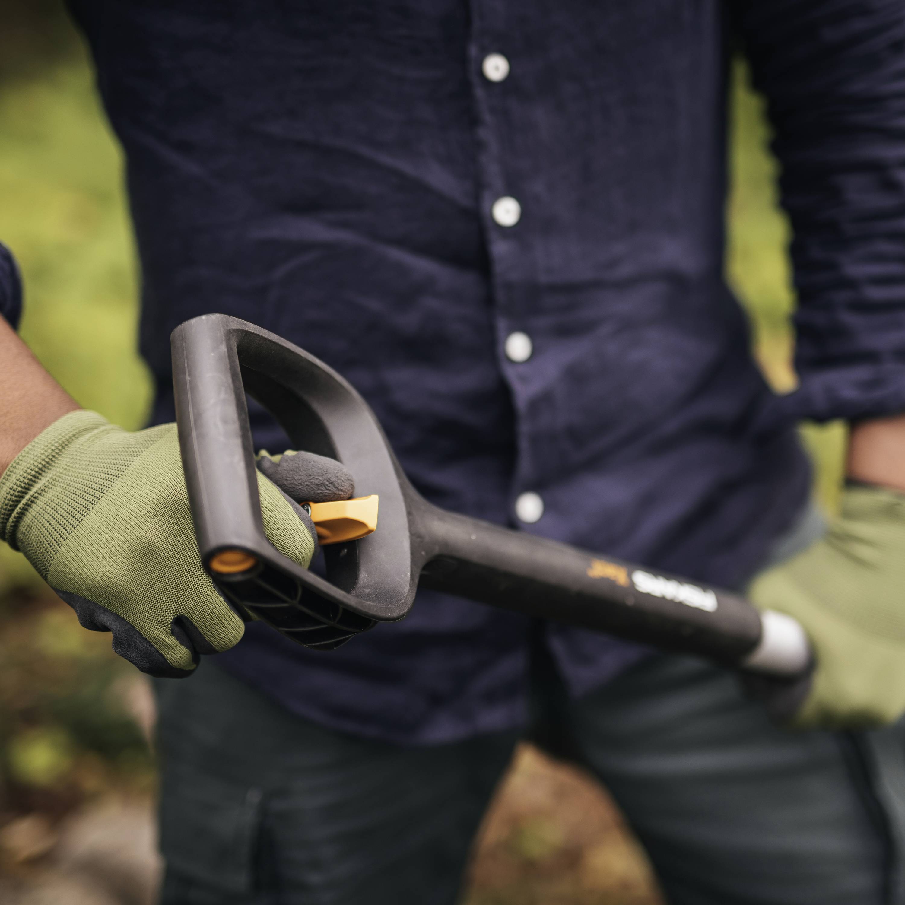 Eine Person in dunkelblauer Kleidung und grünen Handschuhen hält eine Schaufel, bereit für Gartenarbeit.