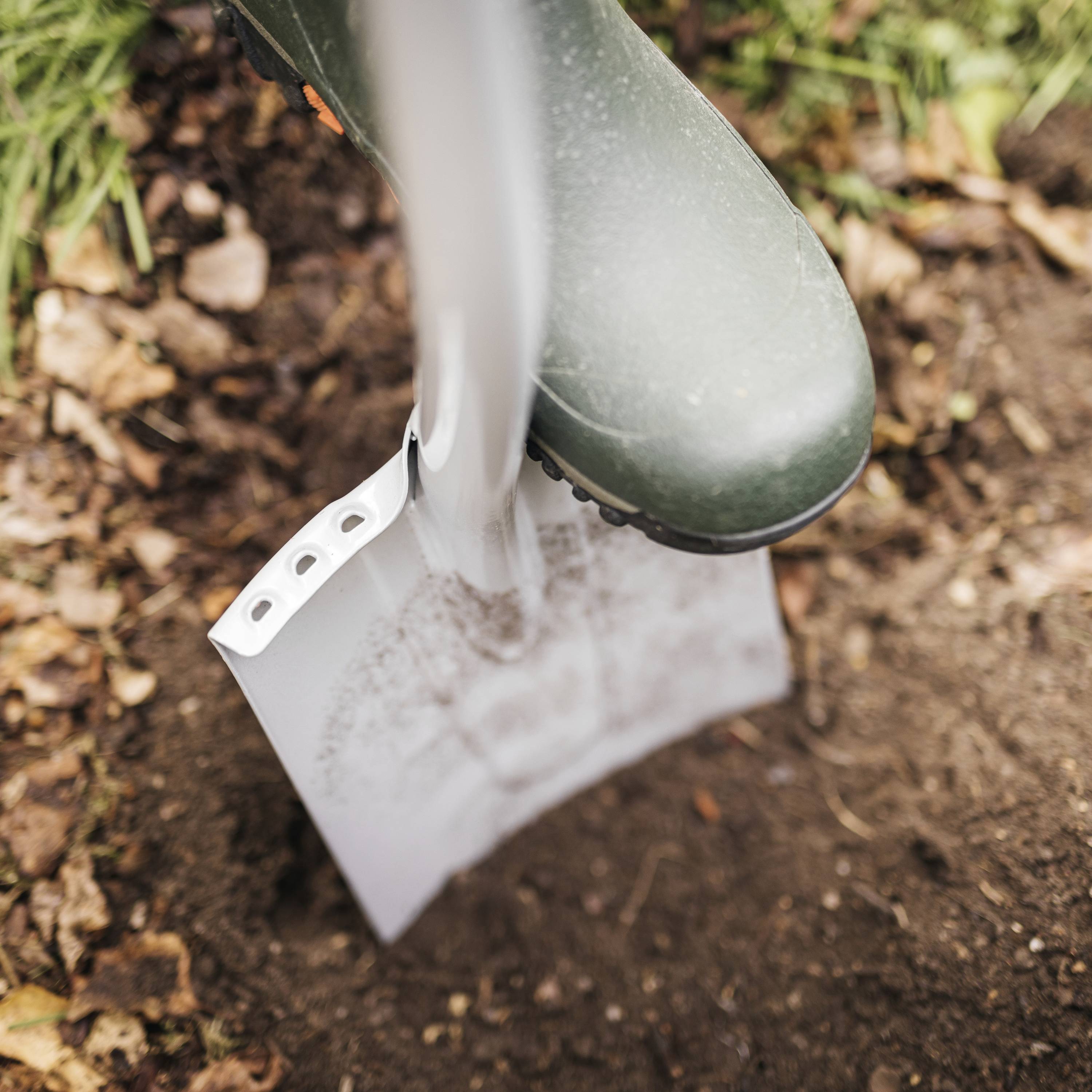 Eine Gummistiefel tragende Person gräbt mit einer Schaufel in Erde. Umgebung: Gras und Laub auf dem Boden.