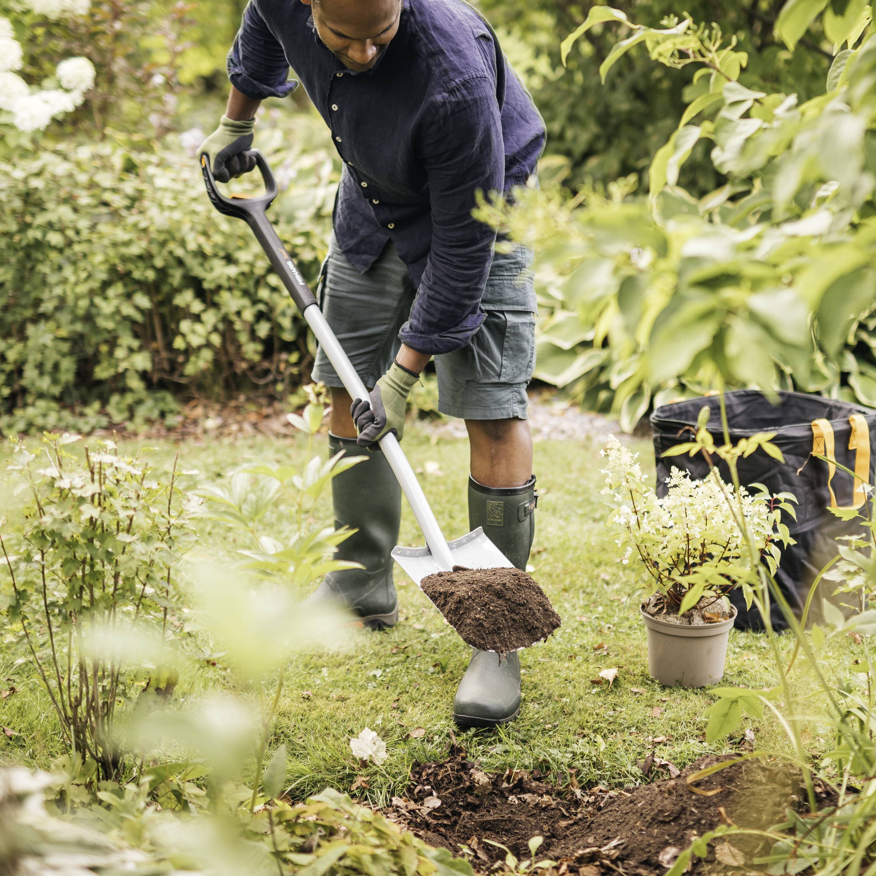 Eine Person in Gummistiefeln gräbt mit einer Schaufel in einem Garten. Um sie herum befinden sich Pflanzen und Büsche.
