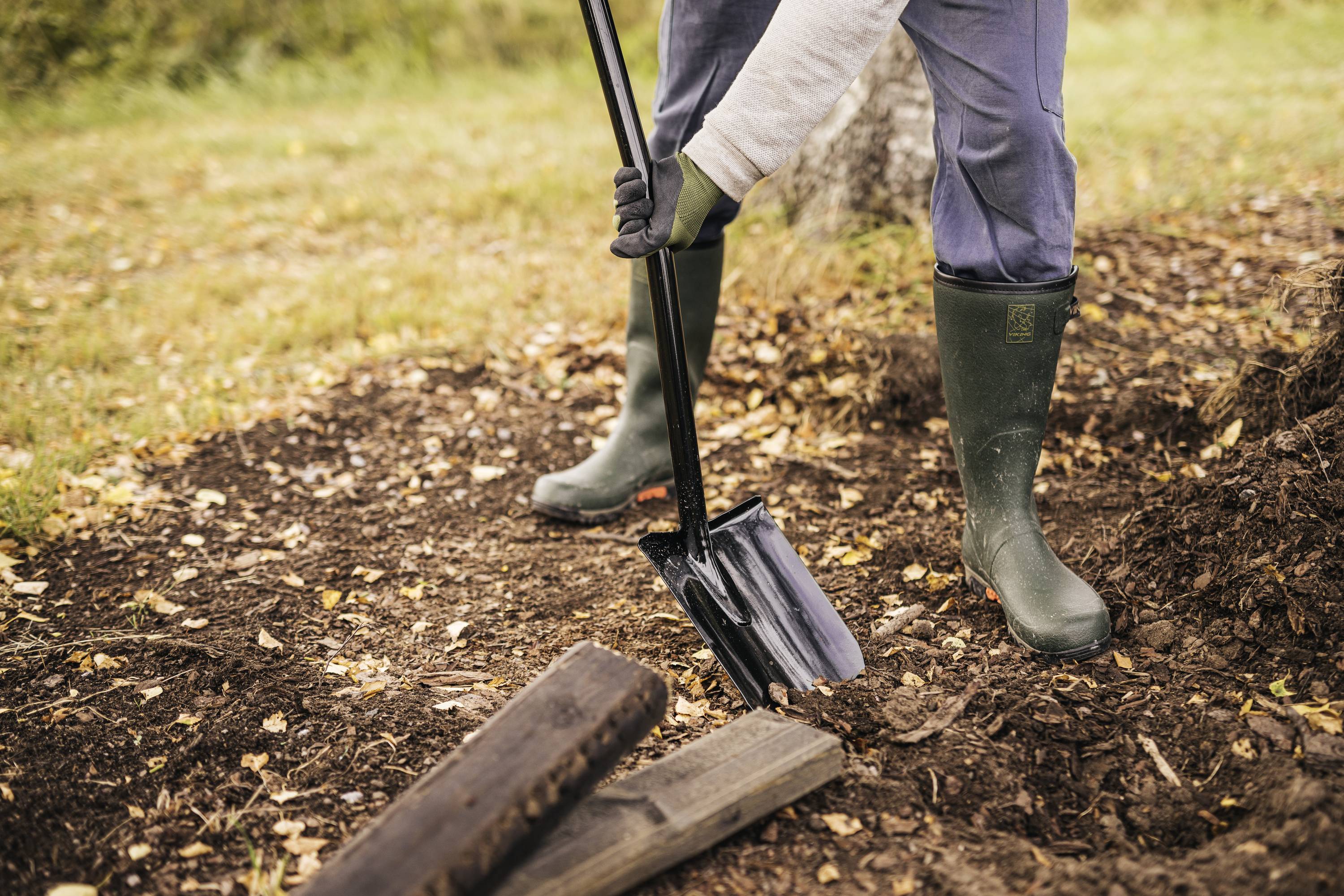 Eine Person mit Gummistiefeln schaufelt Erde in einem Garten. Umgeben von Herbstlaub und Holzstücken auf dem Boden.