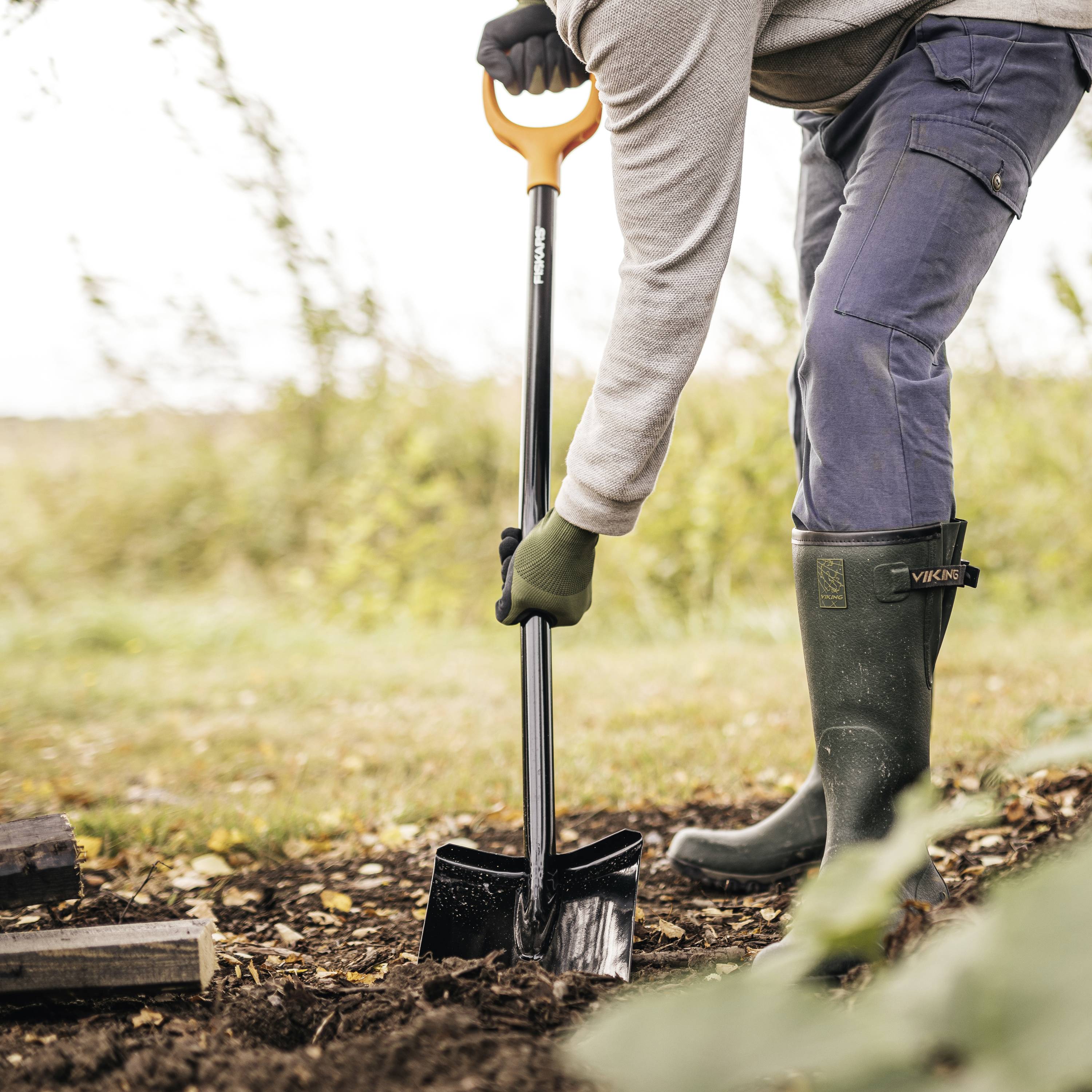 Eine Person in Gartenkleidung hebt Erde mit einer Schaufel. Im Hintergrund ist eine unscharfe Wiese zu sehen.
