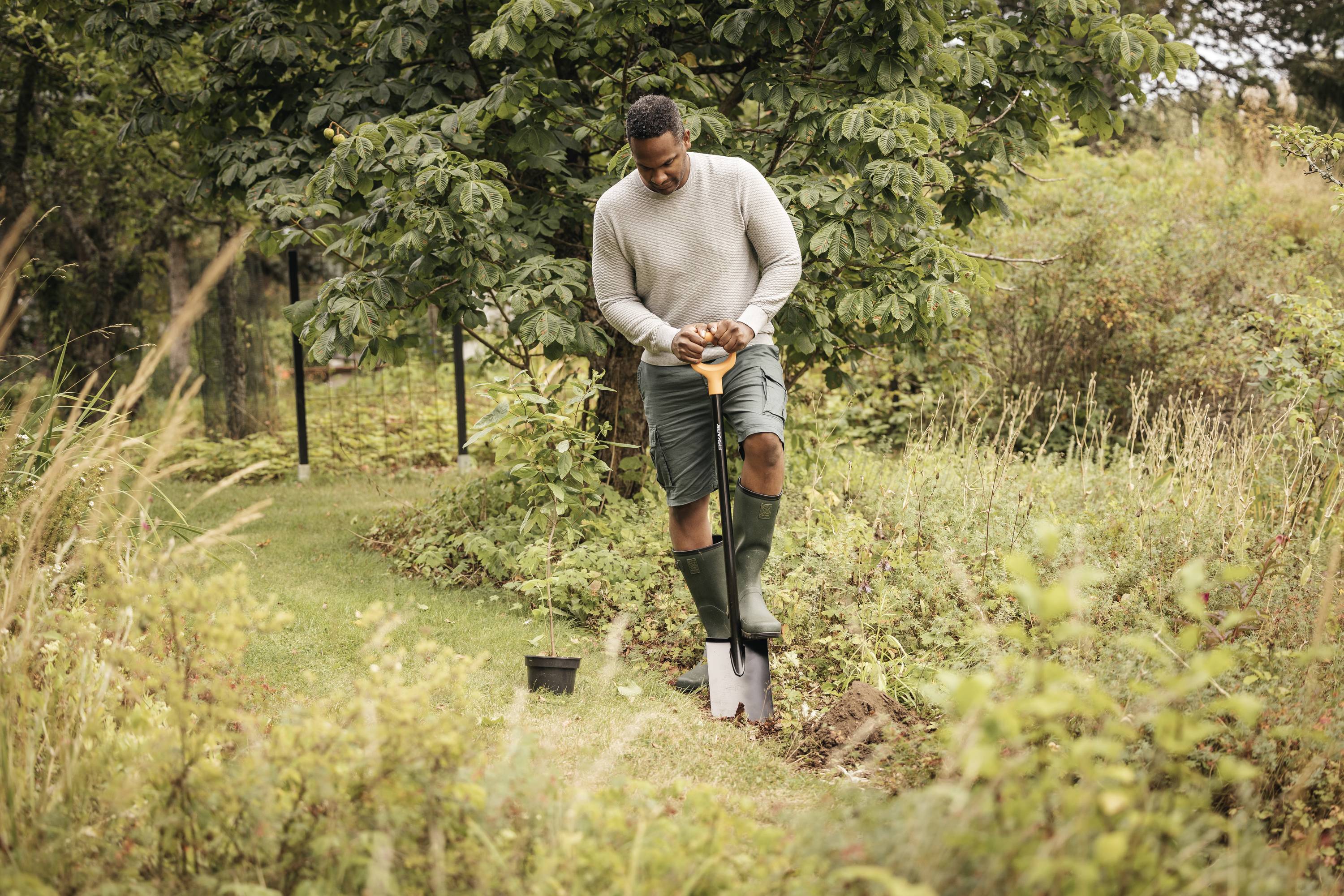 Ein Mann gräbt mit einer Schaufel in einem Garten. Er trägt Gummistiefel und steht in einem Bereich mit grasbewachsenen Pflanzen.