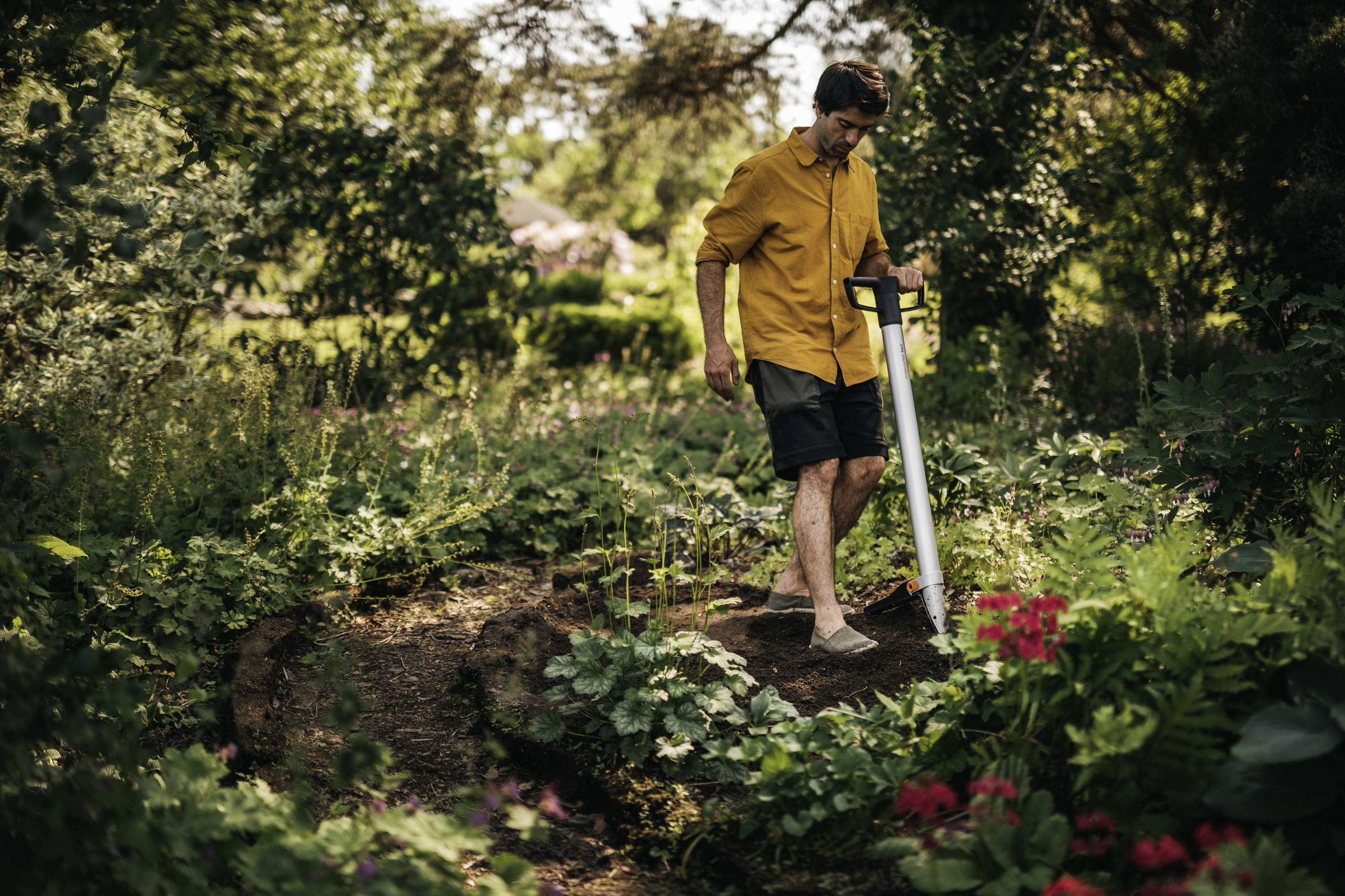 Eine Person in einem gelben Hemd und Shorts geht barfuß durch einen grünen Gartenpfad mit blühenden Blumen und dichter Vegetation.