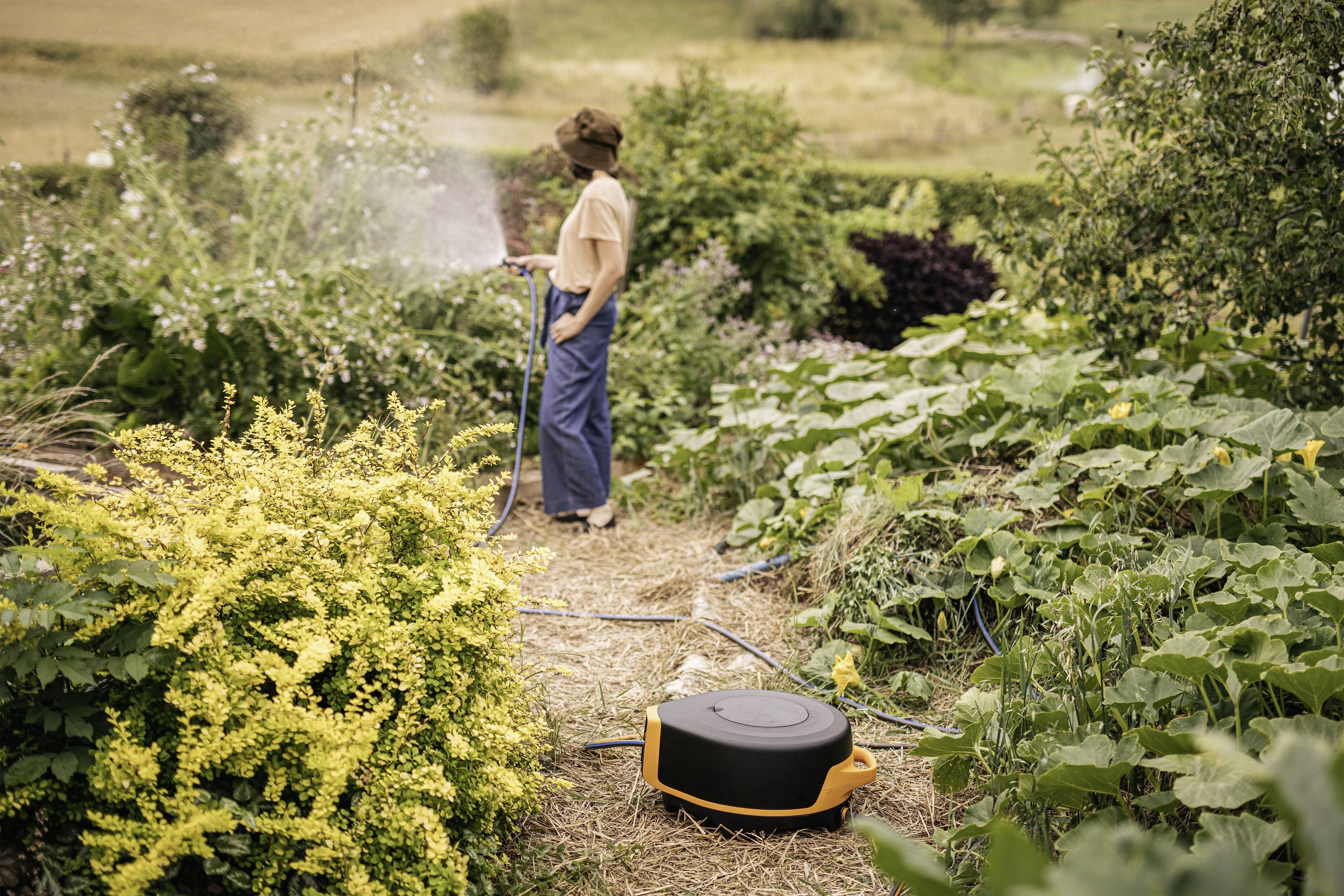 Person gießt mit einem Schlauch den Garten, umgeben von dichtem Grün und Pflanzen; im Vordergrund liegt ein Gartenschlauch-Aufroller.