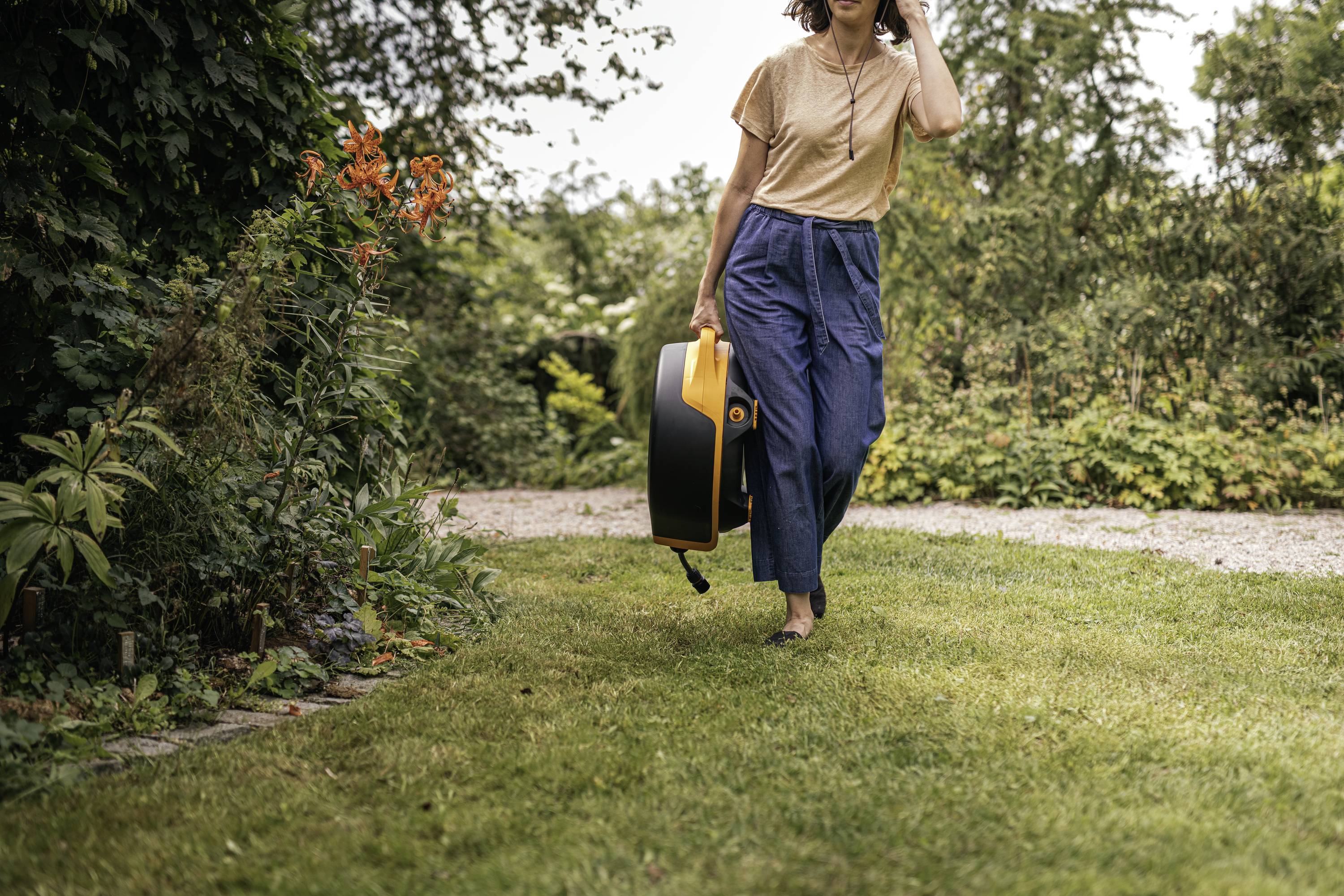 Person im Garten trägt ein Musikinstrument in einem gelben Koffer. Umgebung mit Büschen und Rasen im Hintergrund.