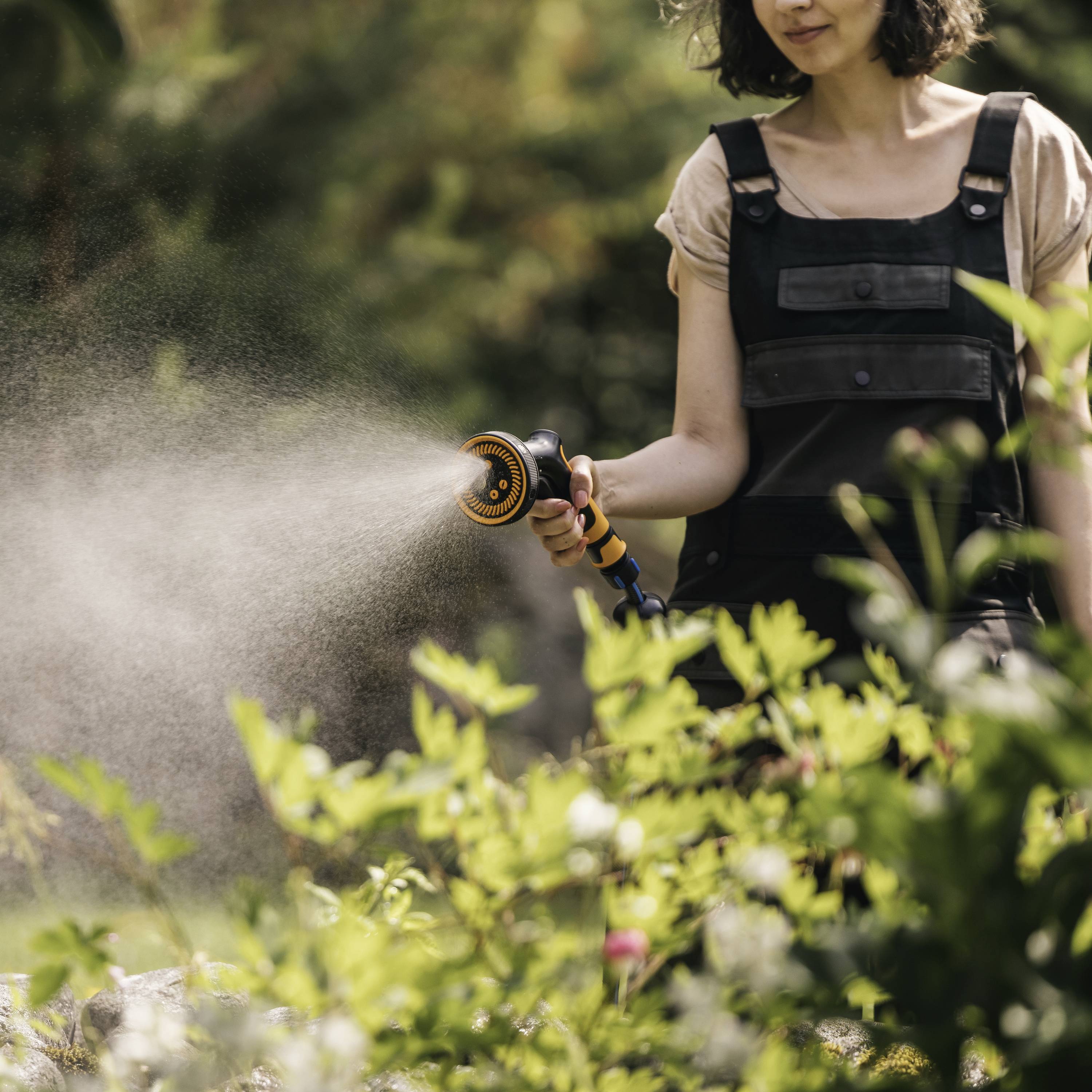 Eine Person gießt tagsüber im Garten Pflanzen mit einem Schlauch, wobei Wassernebel in der Luft schwebt.