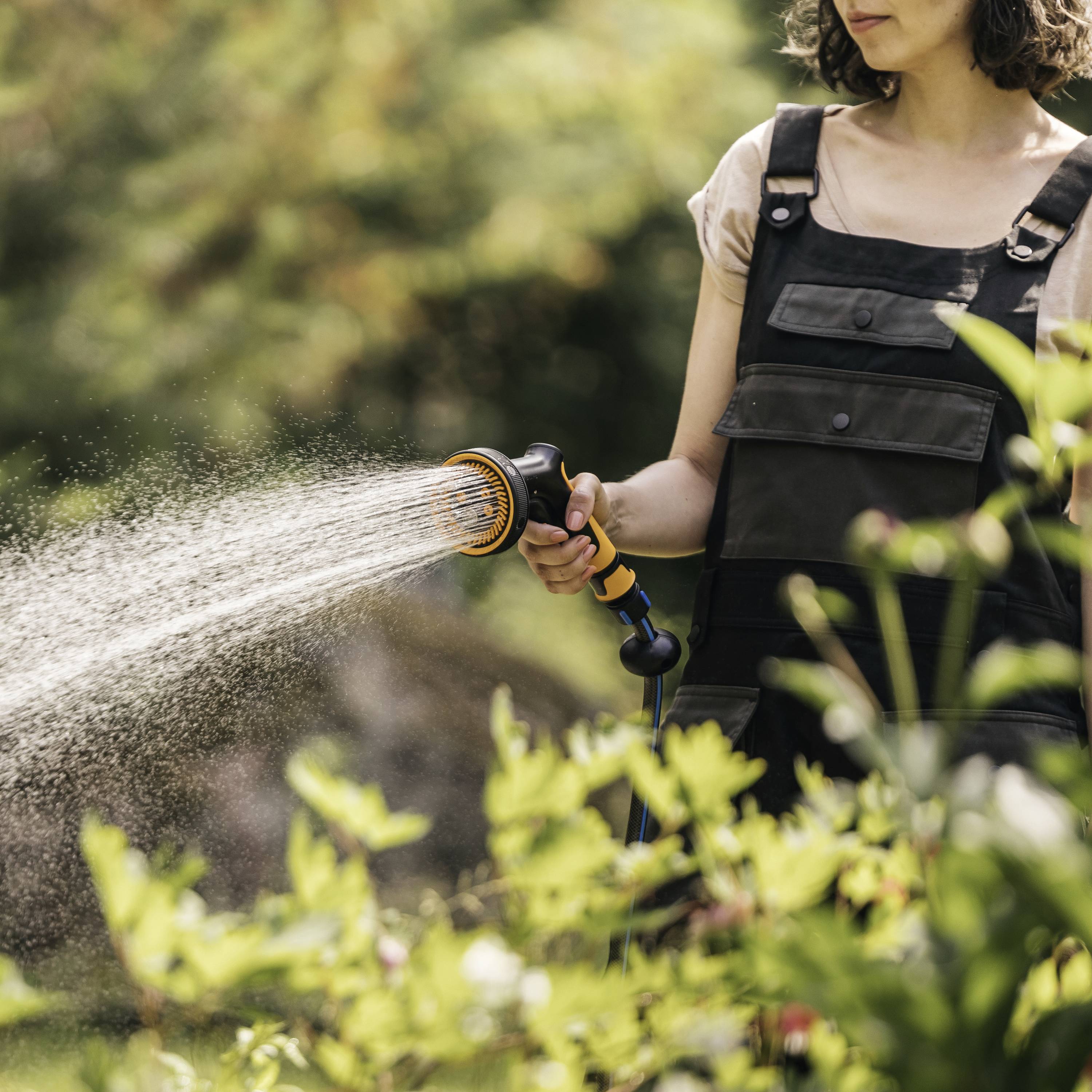 Person sprüht Wasser mit Gartenschlauch auf Pflanzen. Die Person trägt eine Latzhose und steht in einem grünen Gartenbereich.
