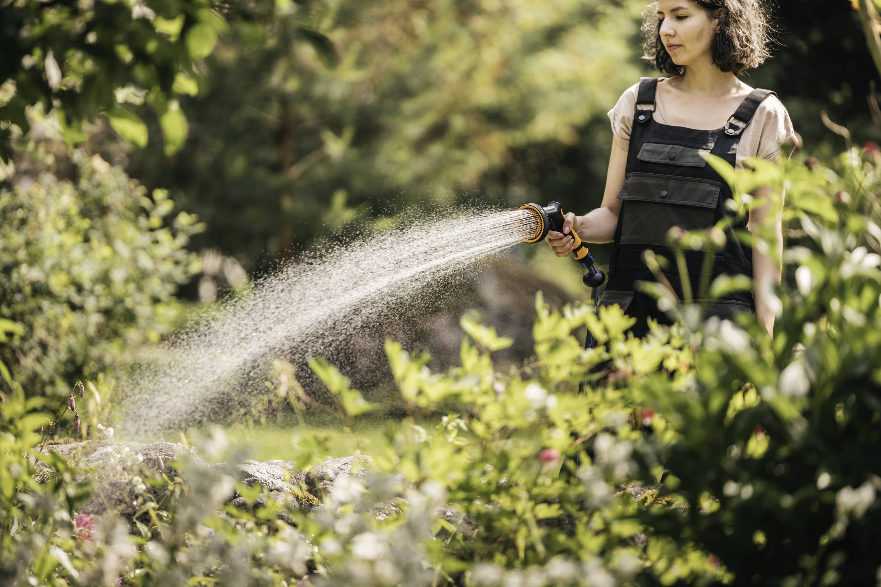 Frau im Garten gießt Pflanzen mit einem Wasserschlauch an einem sonnigen Tag. Hintergrund zeigt üppige grüne Vegetation.