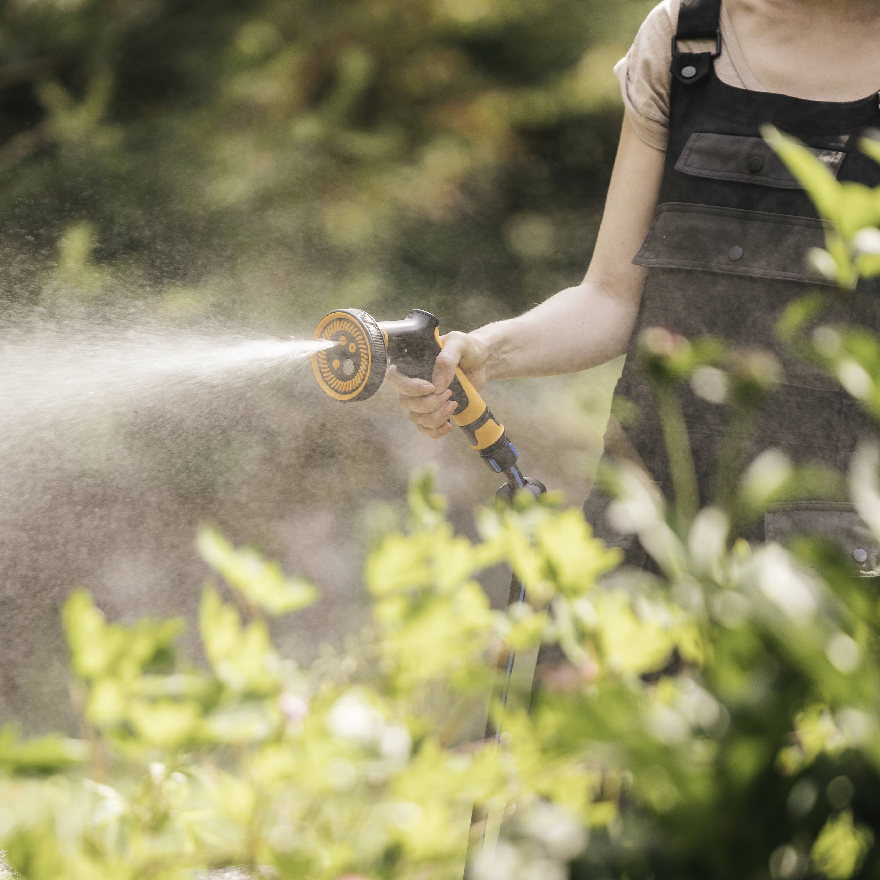 Eine Person mit schwarzem T-Shirt gießt Pflanzen im Garten mit einem Wasserschlauch. Im Vordergrund sind grüne Blätter zu sehen.