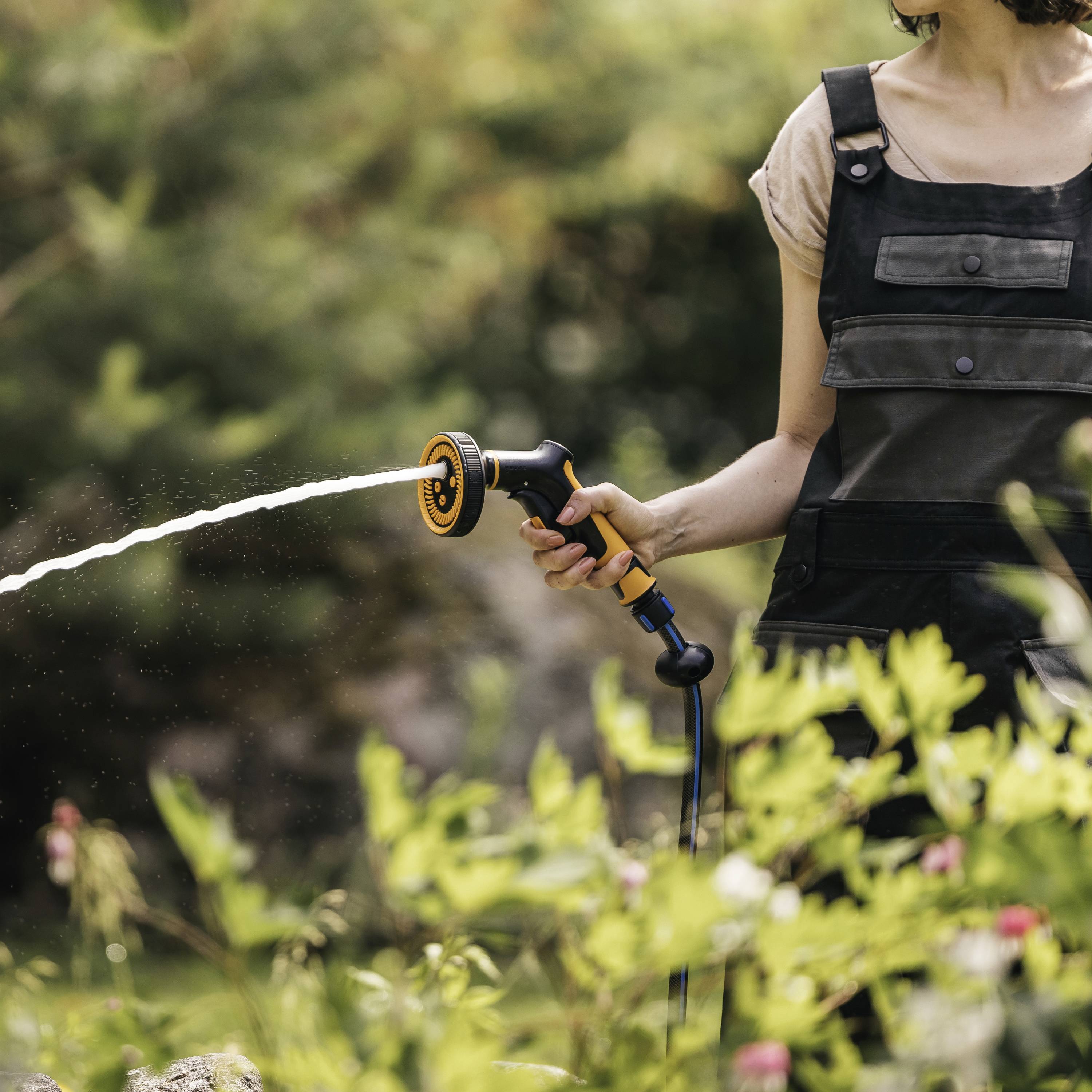 Eine Person in Latzhose gießt Pflanzen im Garten mit einem Wasserschlauch, während die Sonne scheint.