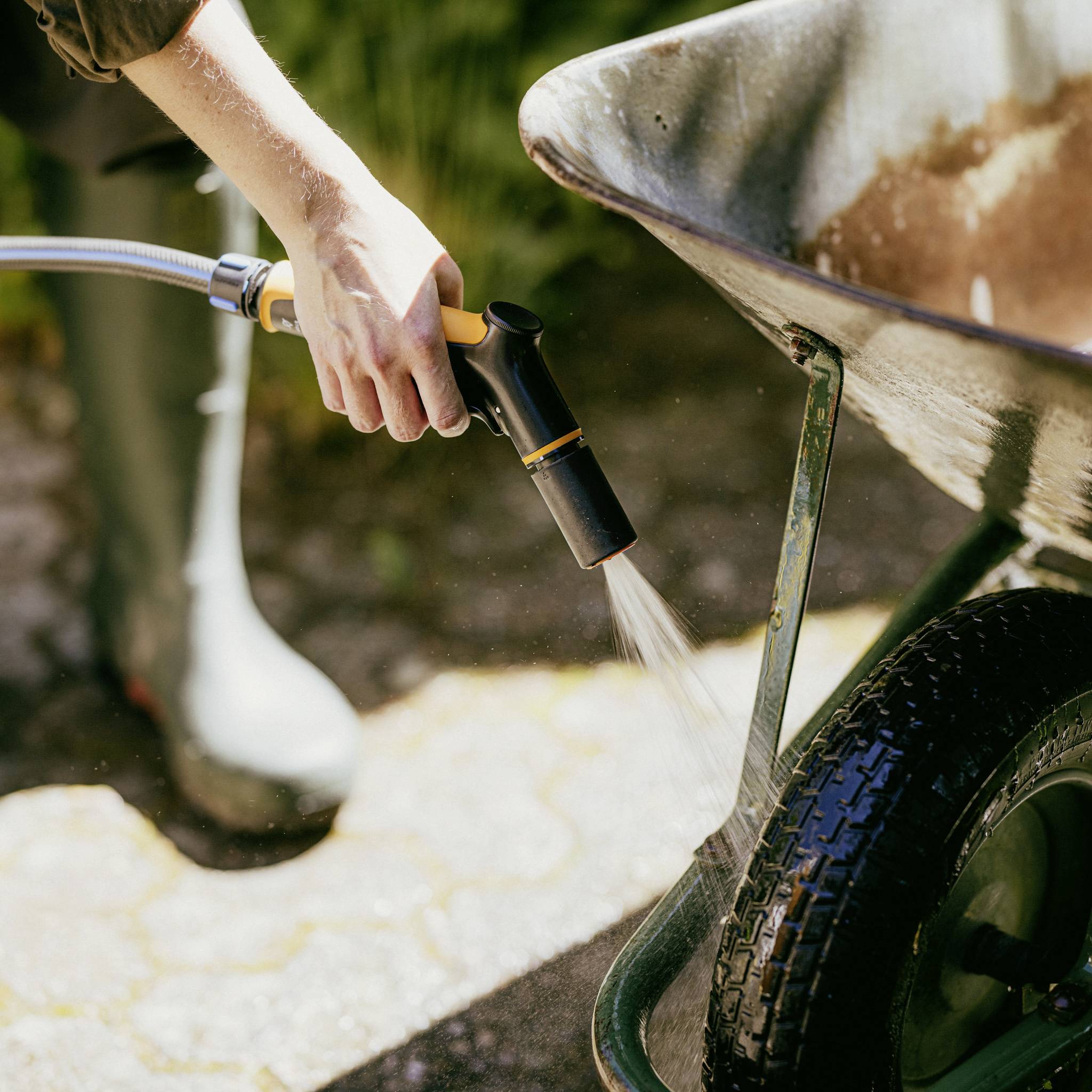 Eine Person spritzt einen Schubkarren mit einem Gartenschlauch ab. Im Vordergrund sind Gummistiefel und nasser Boden zu sehen.