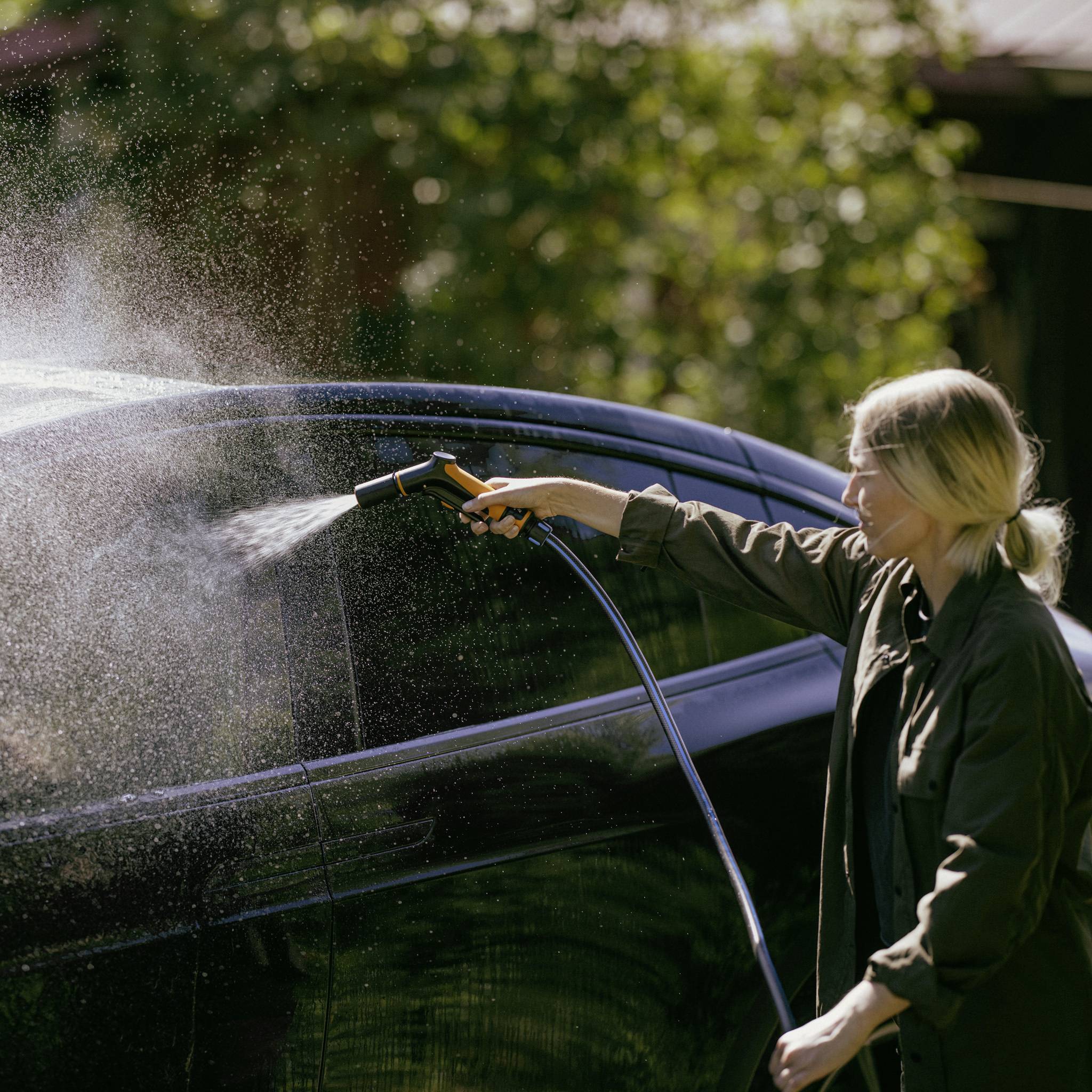 Eine Person wäscht mit einem Wasserschlauch ein Auto, das Sonnenlicht reflektiert. Im Hintergrund sind Bäume zu sehen.