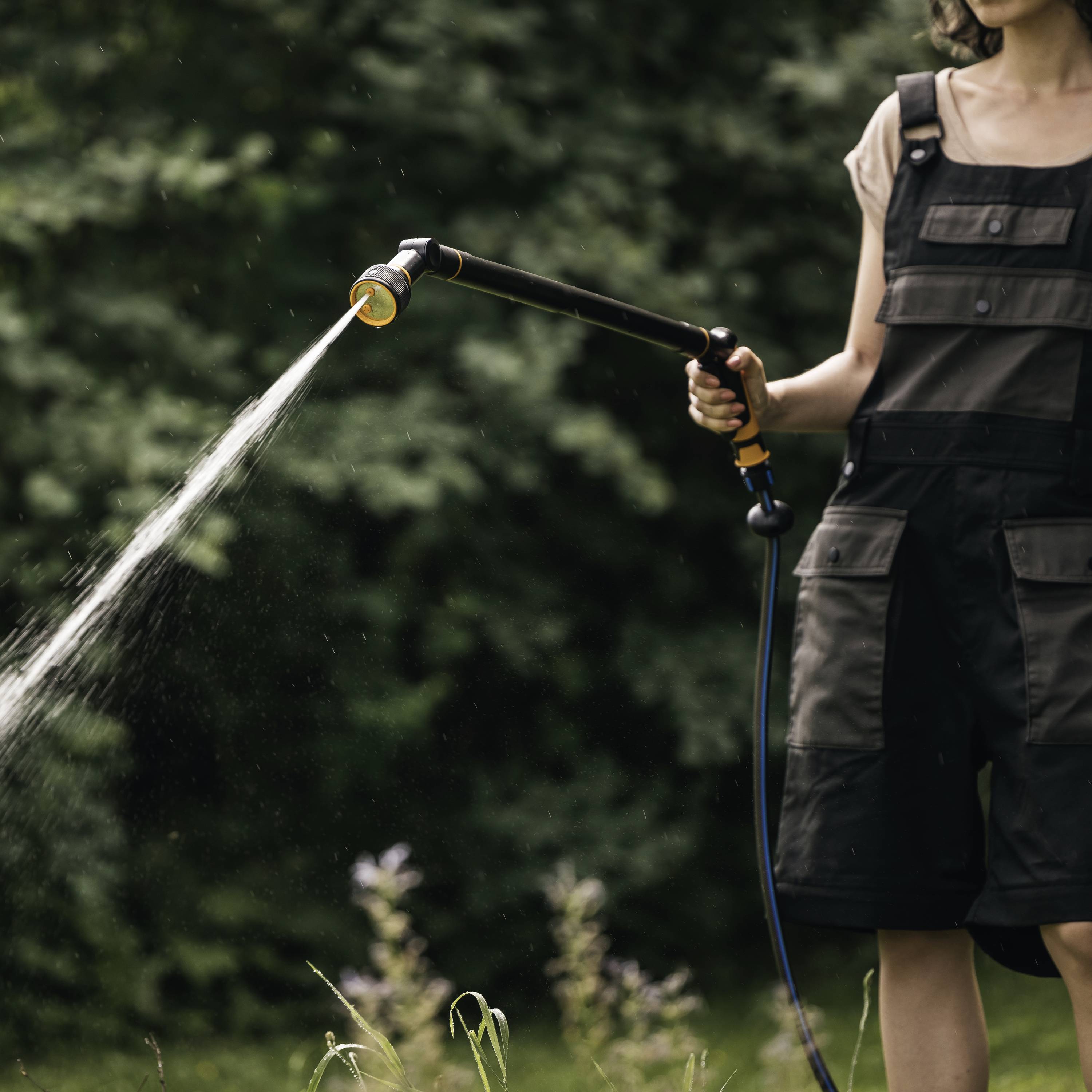 Eine Person in Arbeitskleidung gießt mit einem Wasserschlauch Pflanzen im Freien. Sie steht vor grünen Büschen.