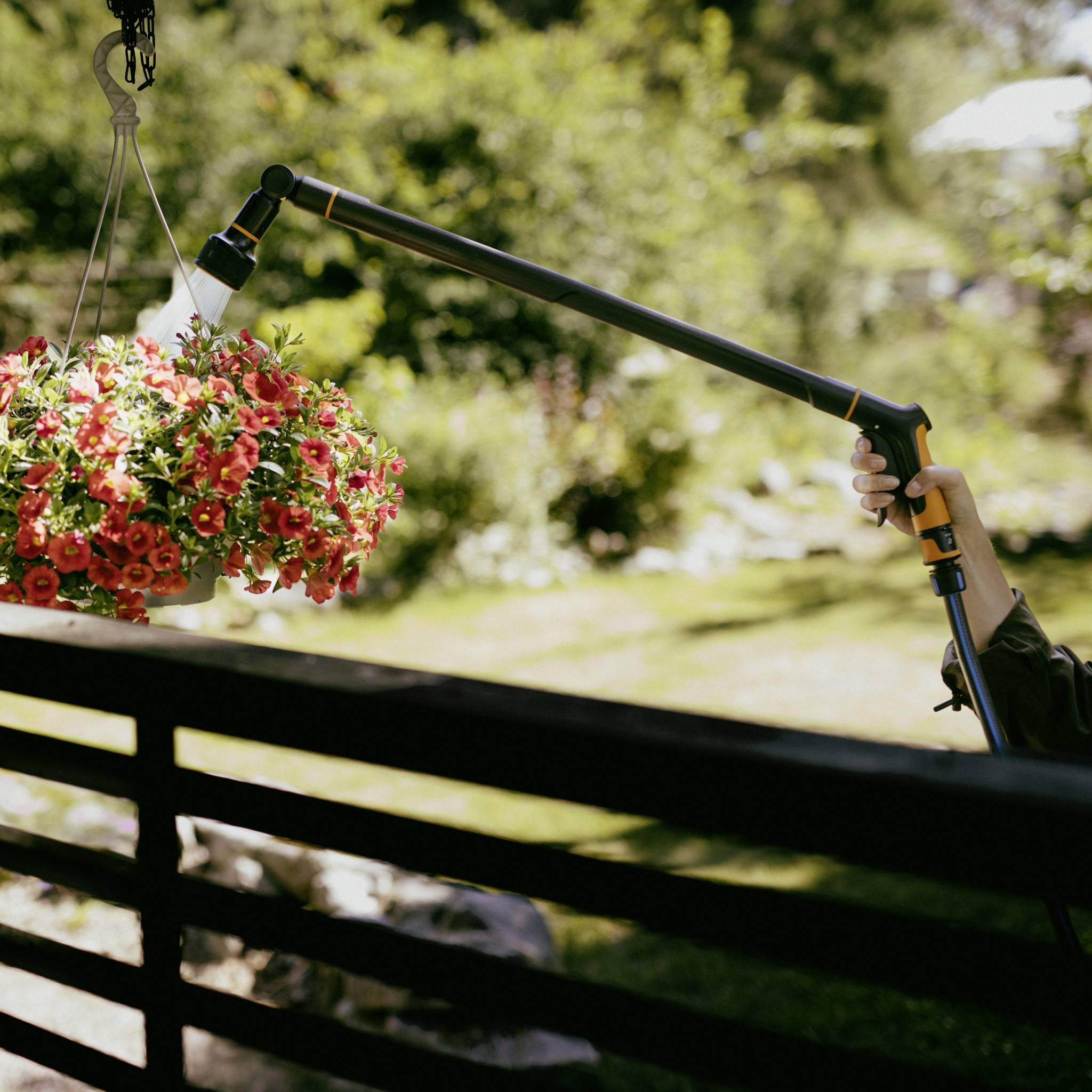Eine Person gießt rote Blumen in einem hängenden Korb mit einer Gießbrause. Im Hintergrund ist ein sonniger Garten zu sehen.