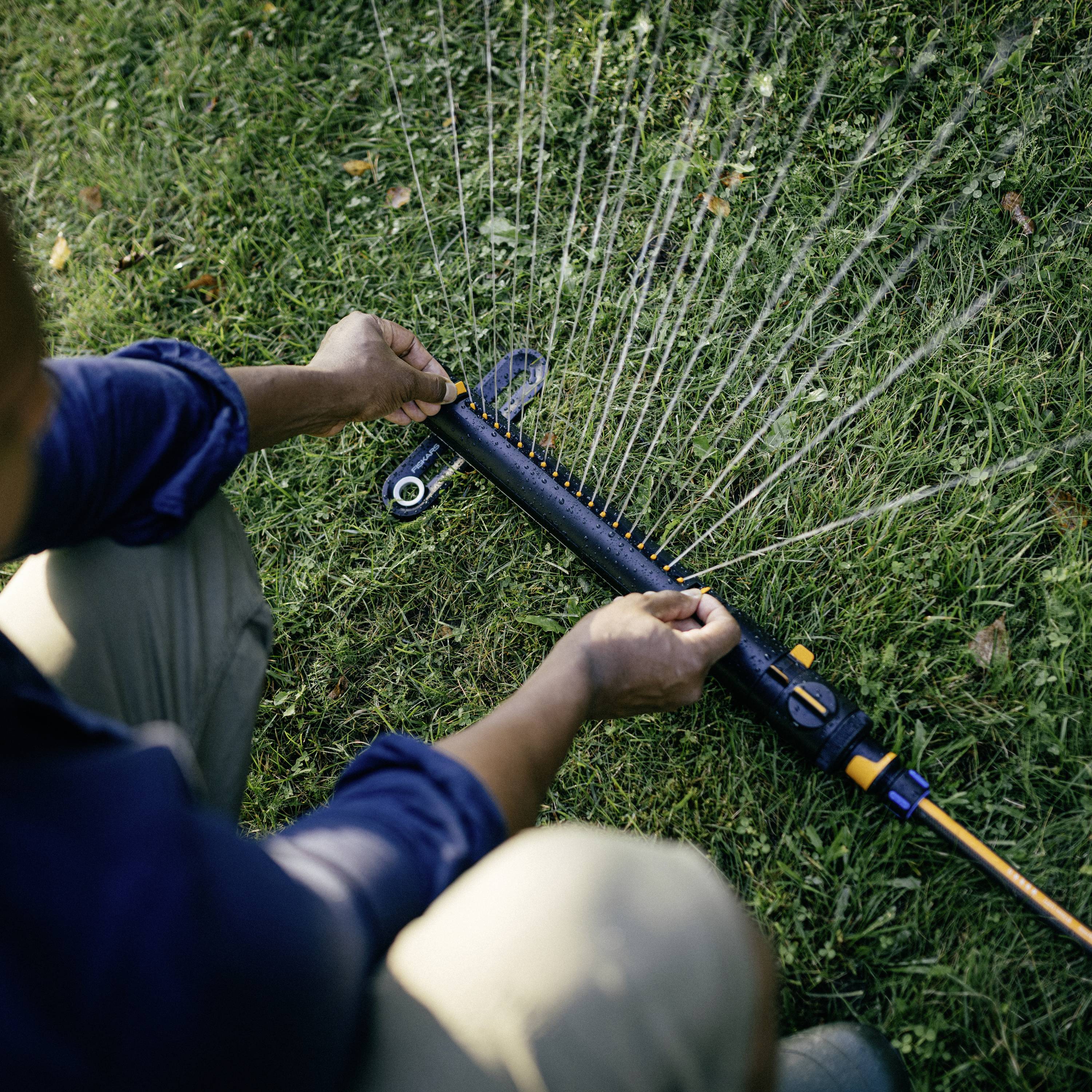Eine Person justiert einen Sprinkler auf einem Rasen. Wasserstrahlen sprühen in einem Bogen, um den Bereich gleichmäßig zu bewässern.