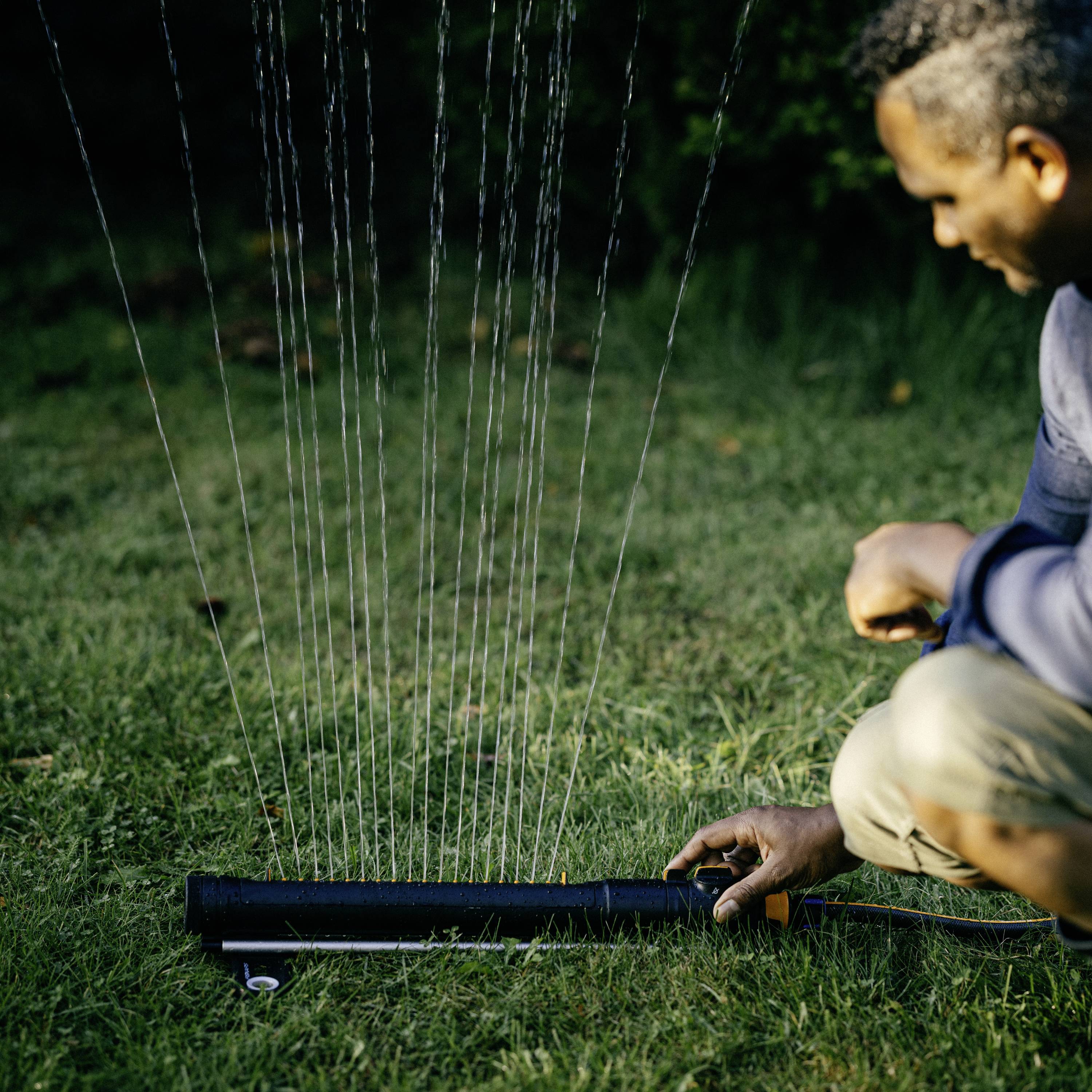Ein Mann kniet auf einem Rasen und hält einen Rasensprenger. Wasserstrahlen sprühen in die Luft.