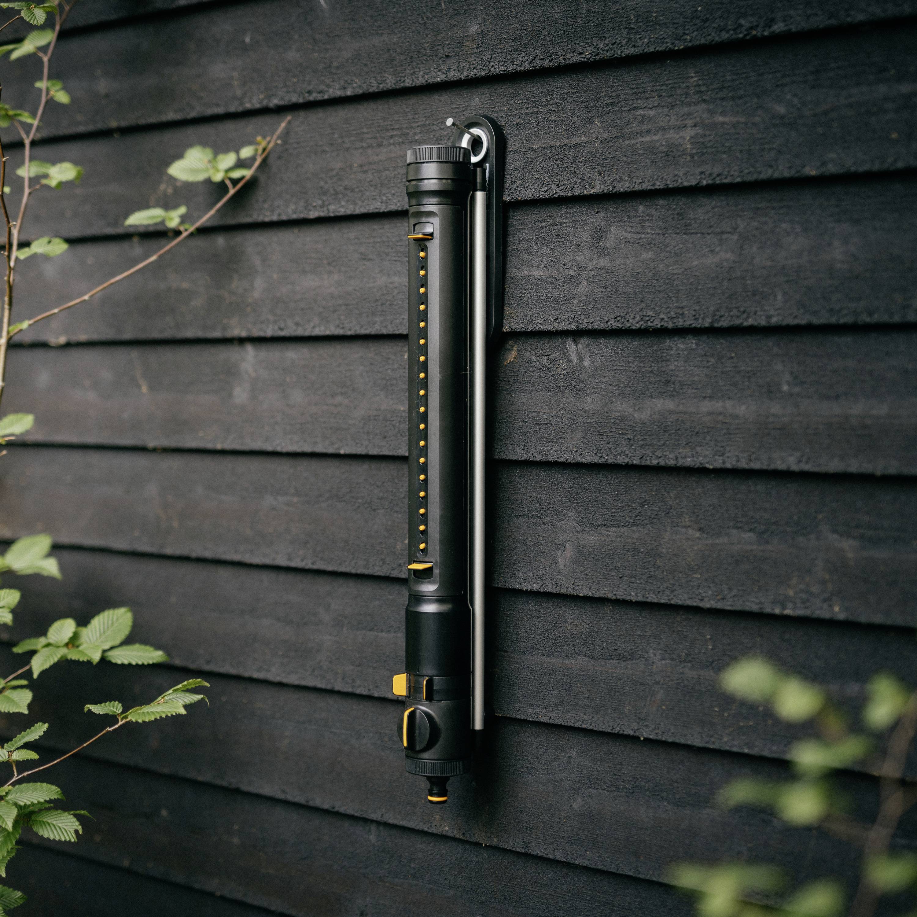 Ein vertikales Wasserthermometer aus Metall, das an einer dunklen Holzwand montiert ist. Pflanzenblätter sind im Vordergrund sichtbar.