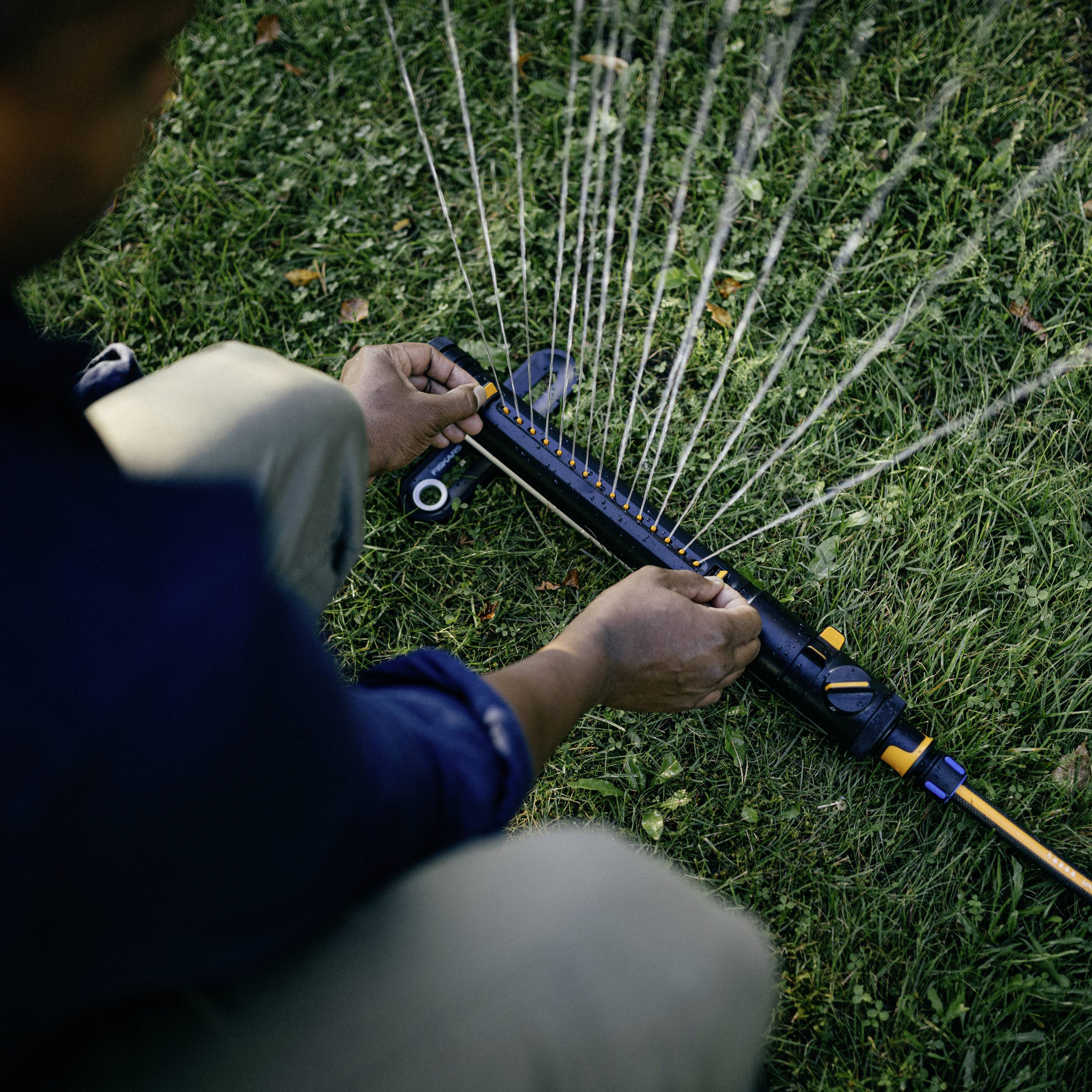 Eine Person kniet im Gras und justiert einen Rasensprenger, der Wasserstrahlen in verschiedenen Winkeln versprüht.