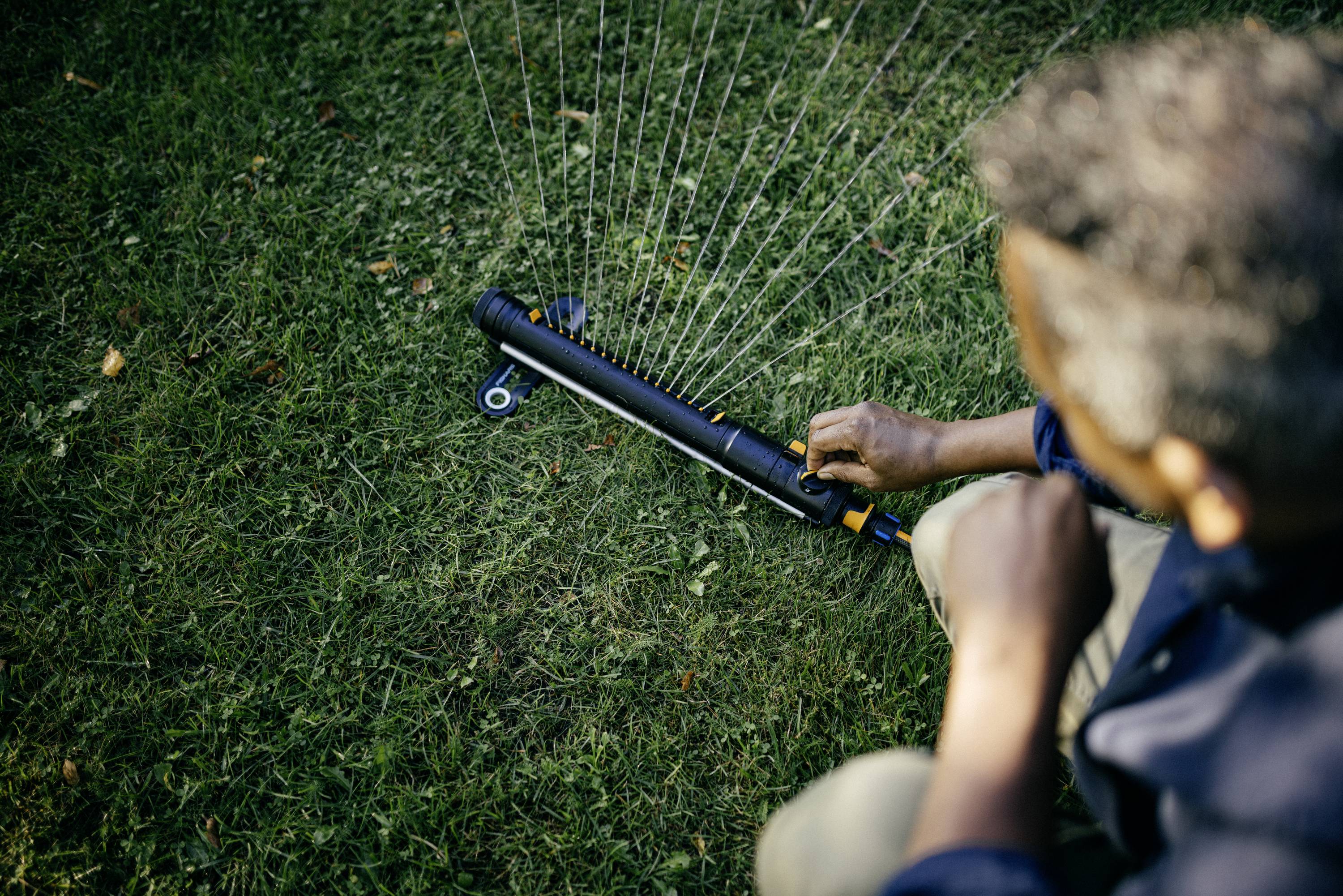 Person stellt auf einer Wiese einen Rasensprinkler ein. Hände bedienen die Einstellung des Sprinklers für gleichmäßige Bewässerung.