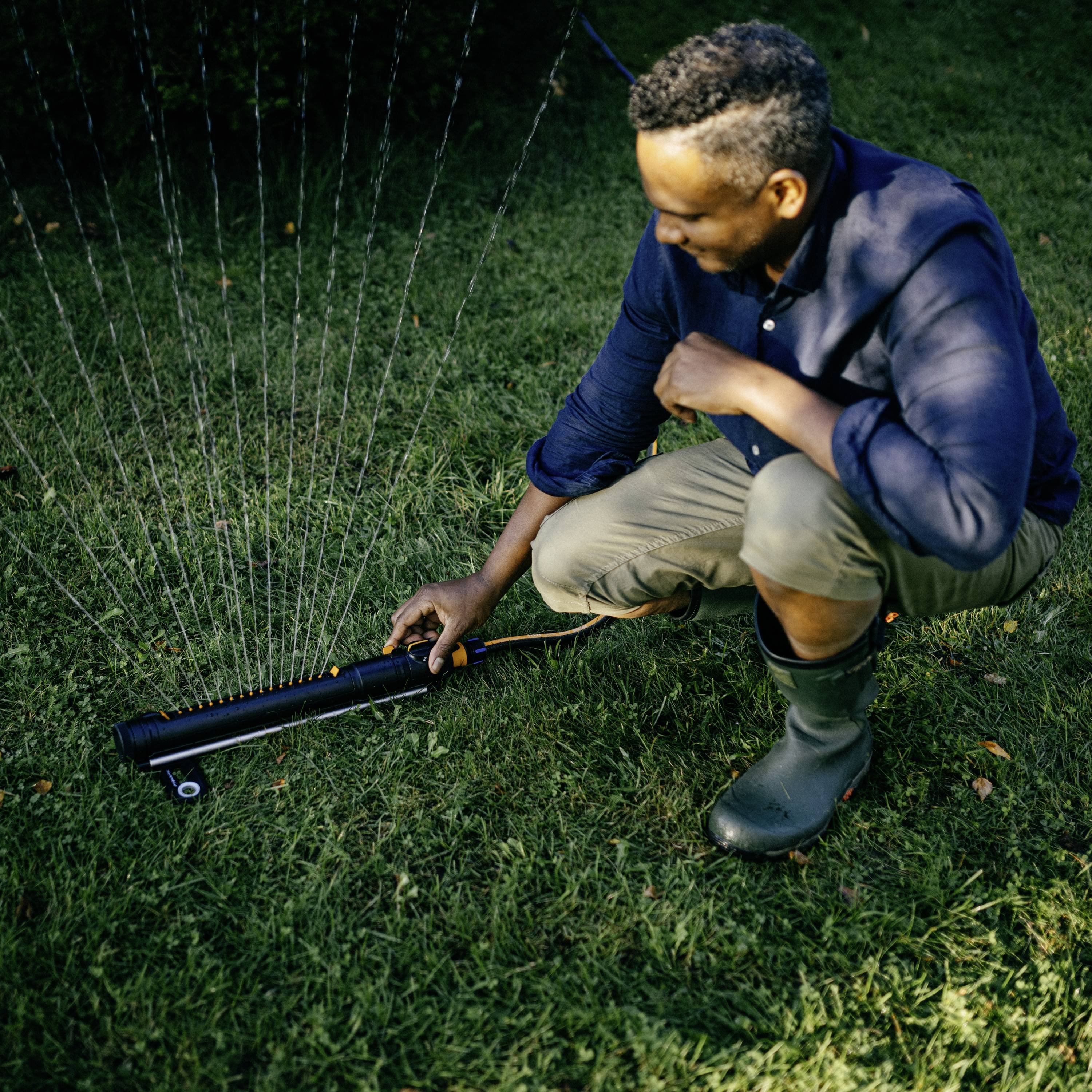 Ein Mann kniet auf einem Rasen und justiert einen Rasensprenger. Es ist tagsüber, das Gras ist grün und dicht.