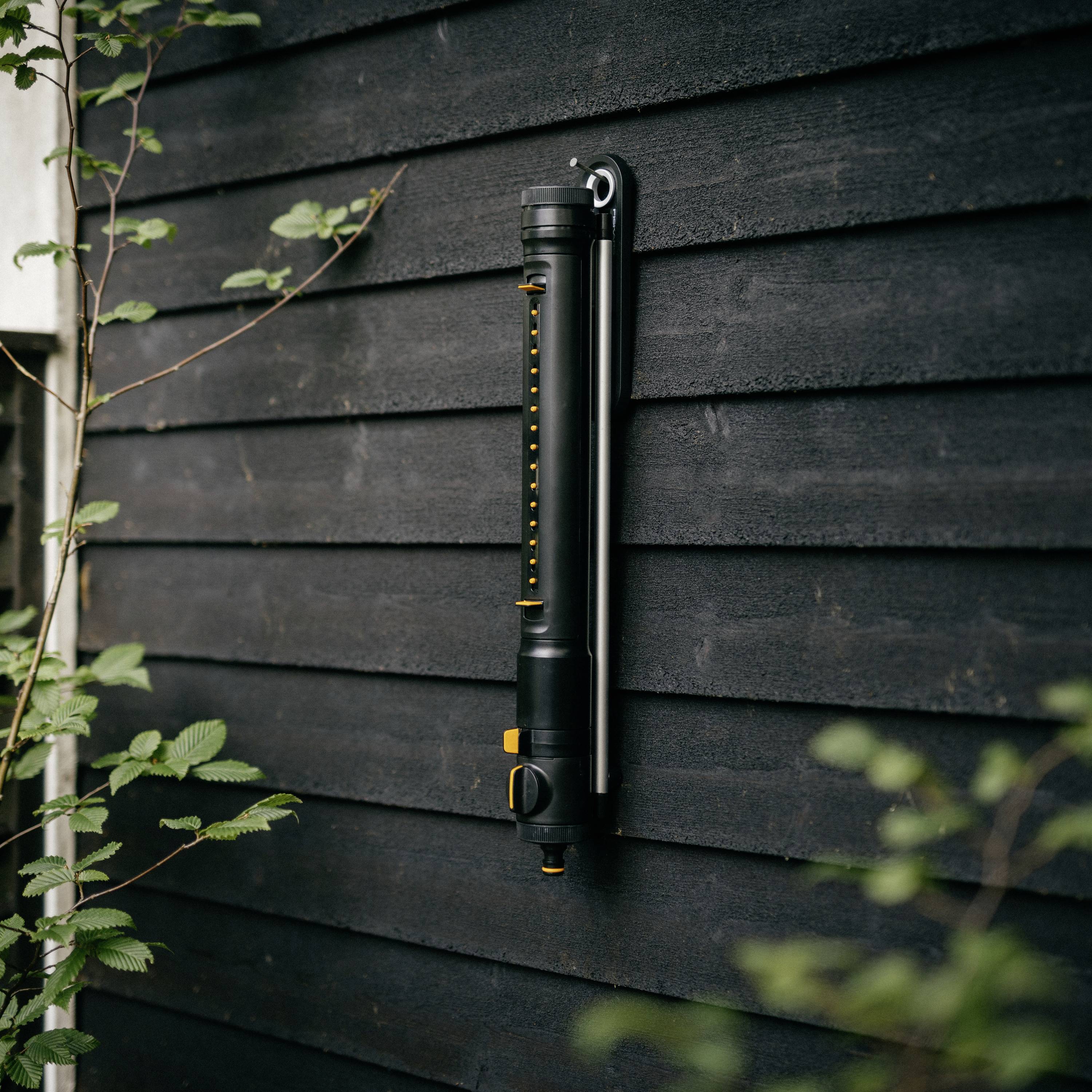Ein schwarzer Gartensprinkler ist an einer dunklen Holzfassade montiert. Pflanzen sind daneben sichtbar.