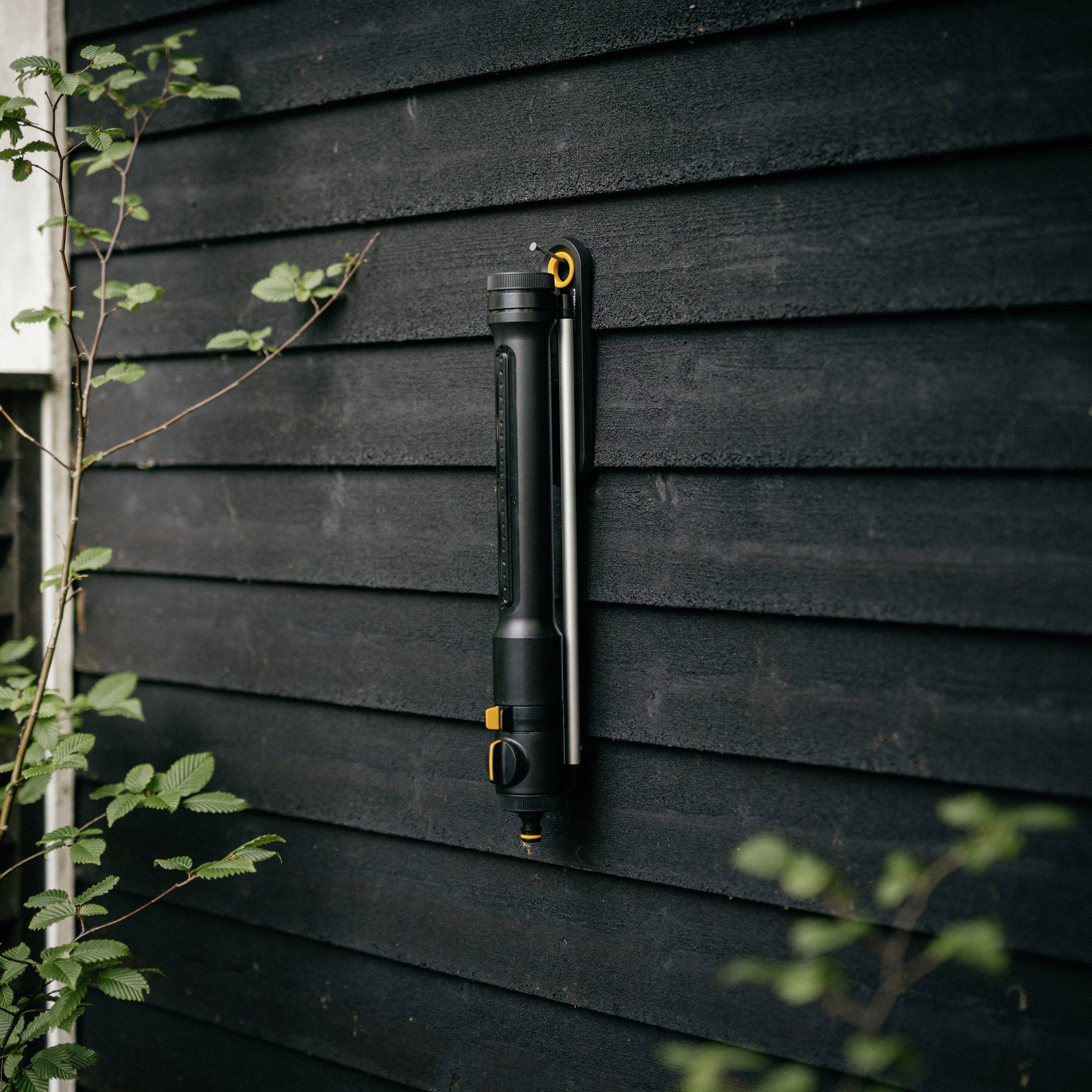 Eine schwarze Wasserpistole mit orangenen Akzenten hängt an einer dunklen Holzlattenwand. Pflanzen sind im Vordergrund zu sehen.