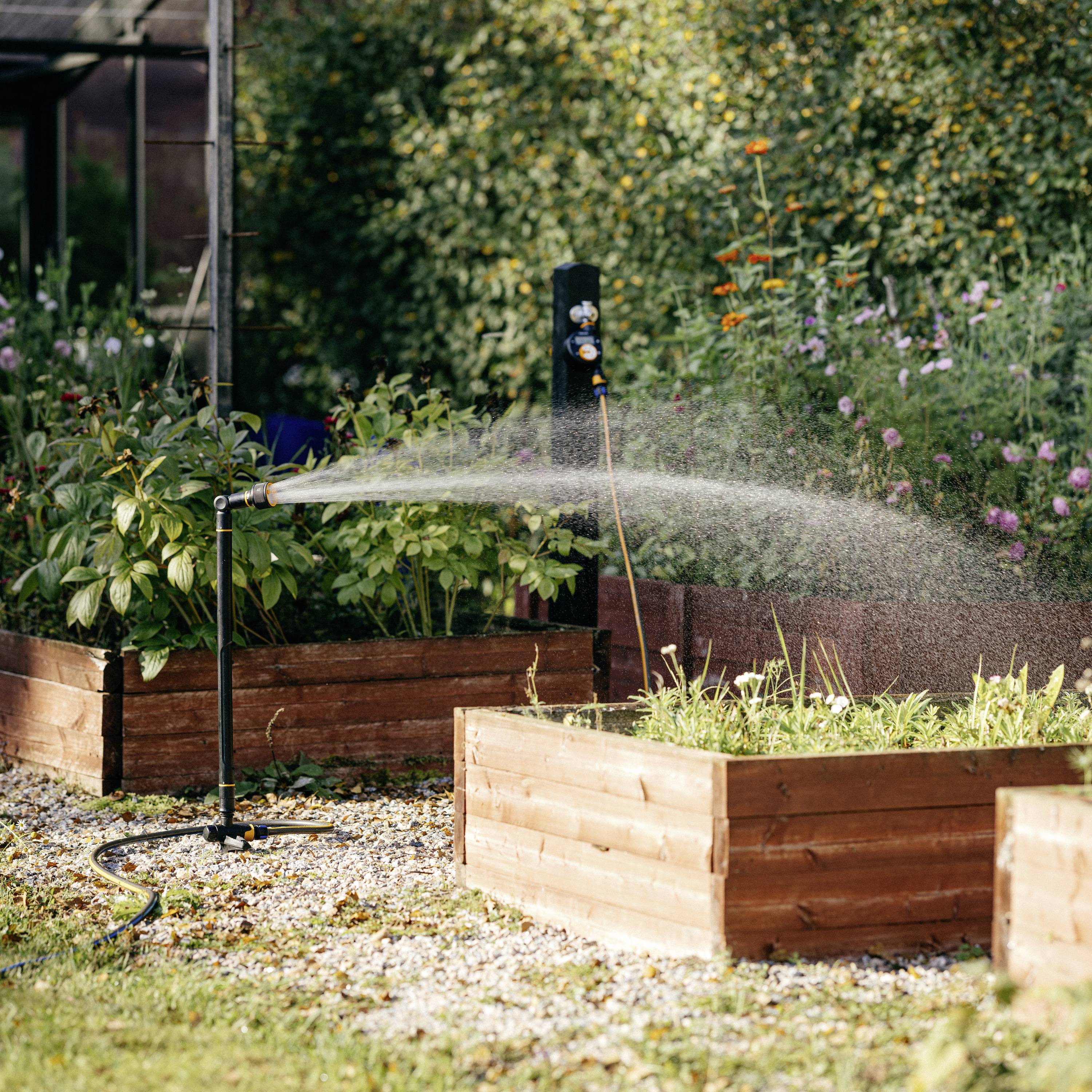Ein automatischer Sprinkler bewässert Beete mit blühenden Pflanzen in einem sonnigen Garten, umgeben von Kieselsteinen.