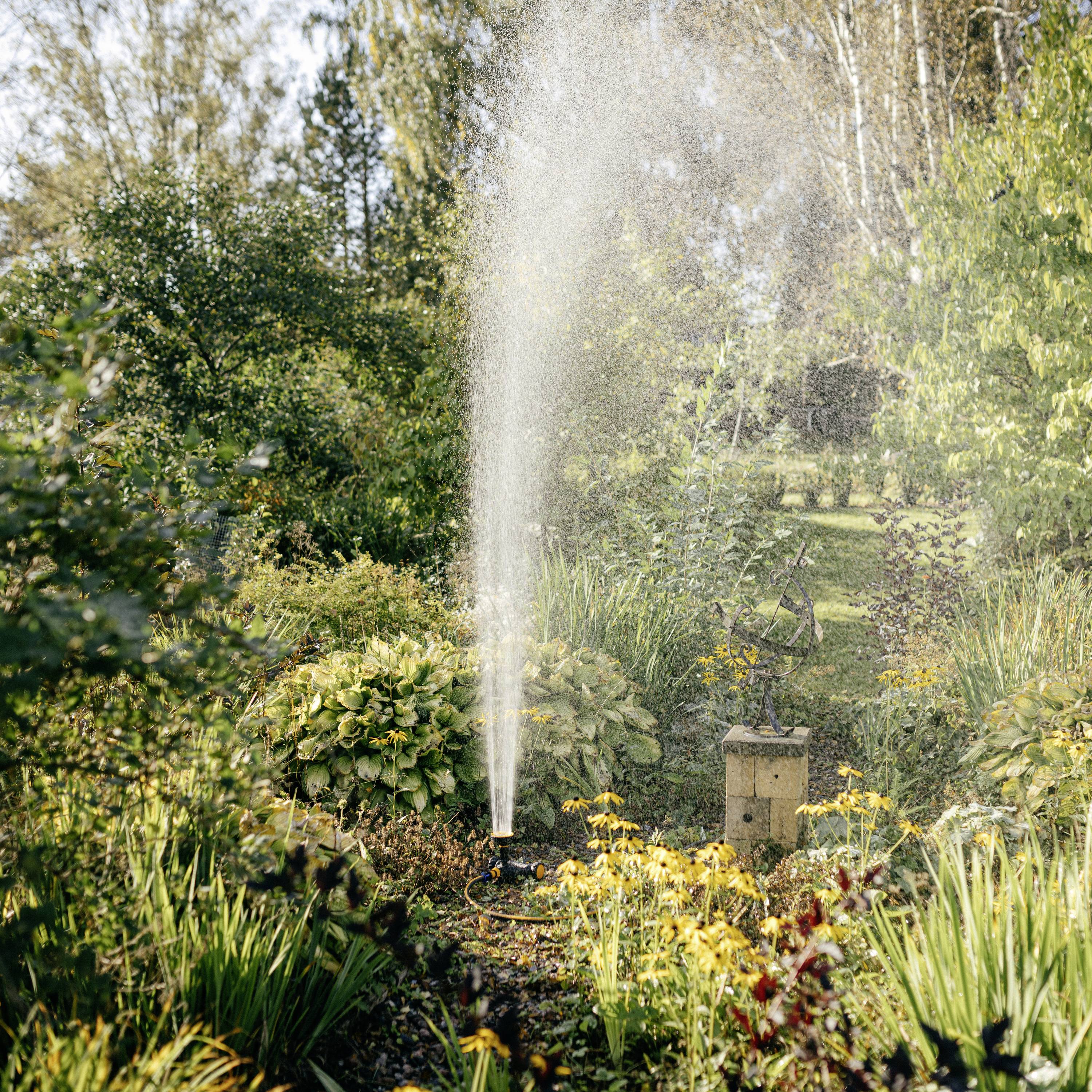 Ein Garten mit blühenden Pflanzen wird von einem an der Seite aufgestellten Sprinkler bewässert, umgeben von Bäumen im Hintergrund.