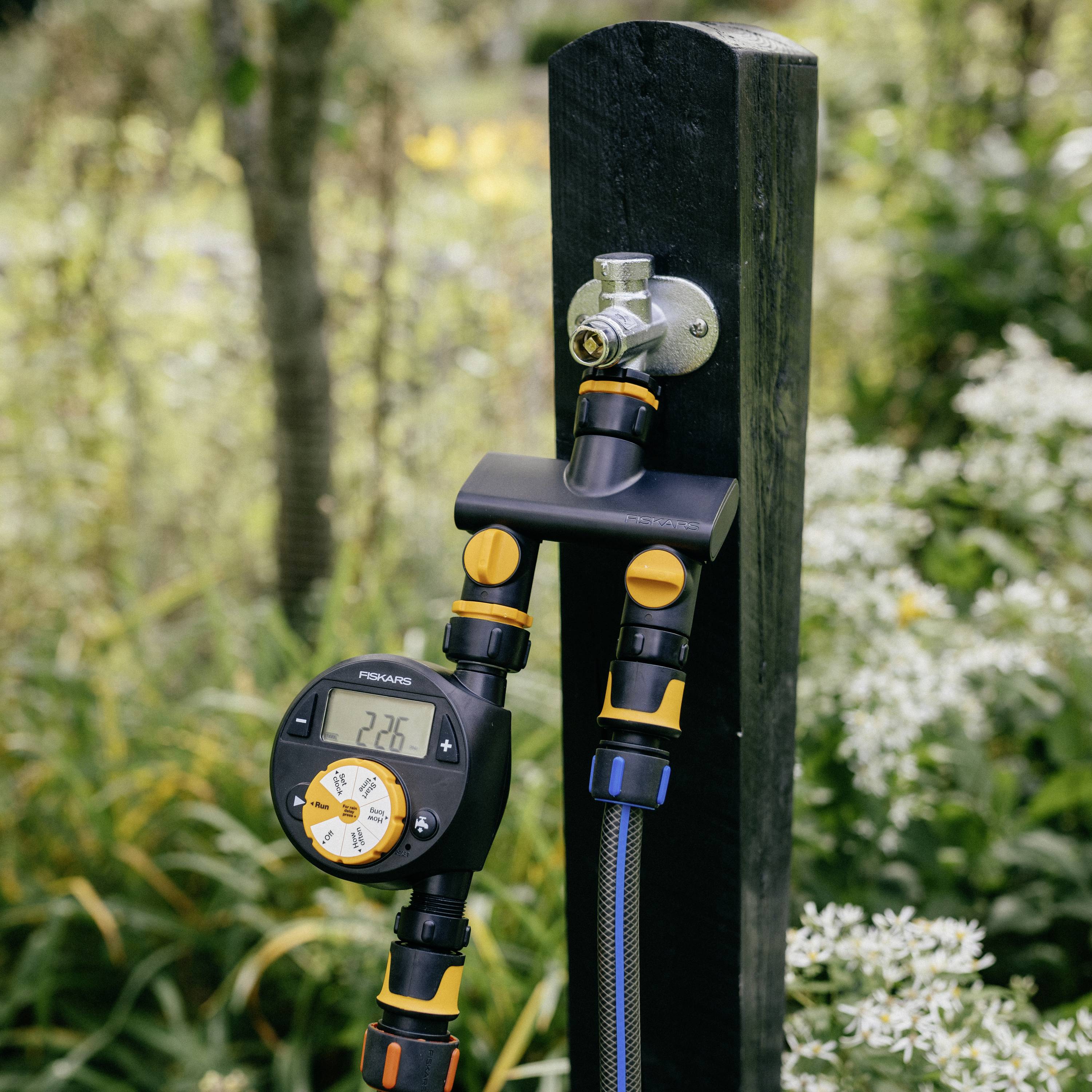 Bewässerungssteuerung an einem Pfosten befestigt, mit Schlauch und digitalem Timer, umgeben von grünen Pflanzen.