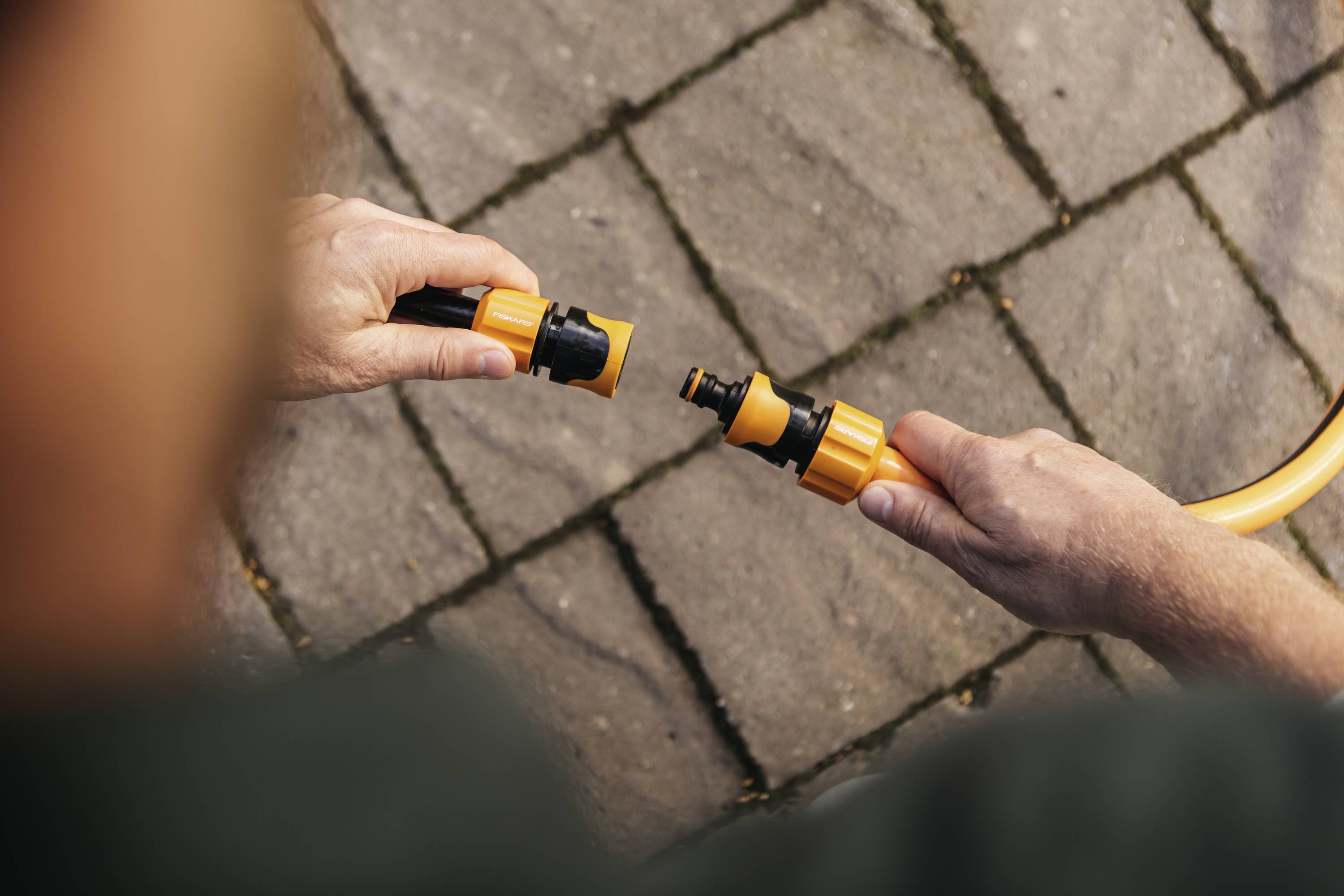 Eine Person verbindet zwei Teile eines Gartenschlauchs mit Steckverbindern auf einem gepflasterten Boden.