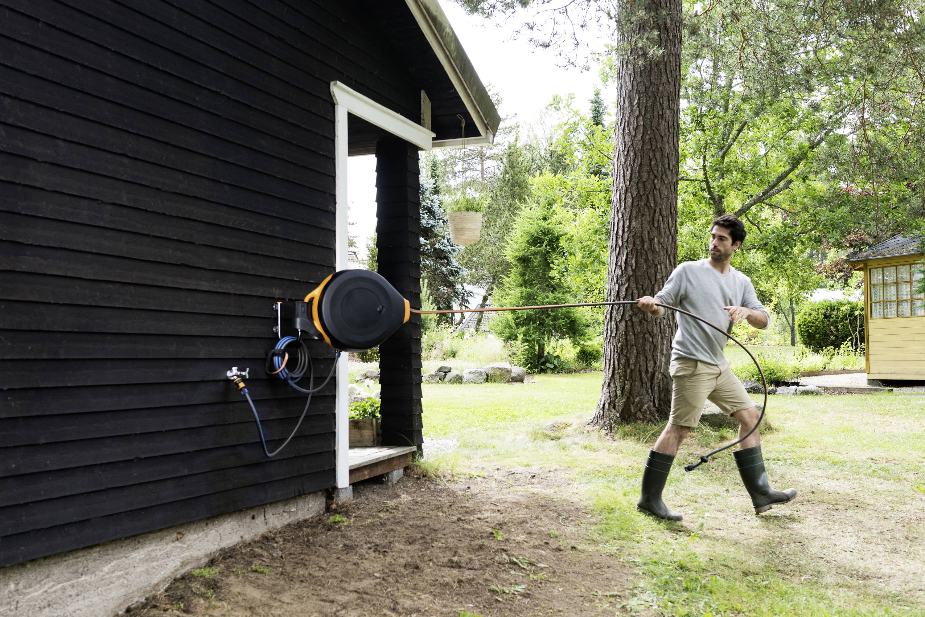 Ein Mann zieht einen Gartenschlauch aus einer Wandhalterung an einem dunklen Holzhaus in einem grünen Gartenbereich.