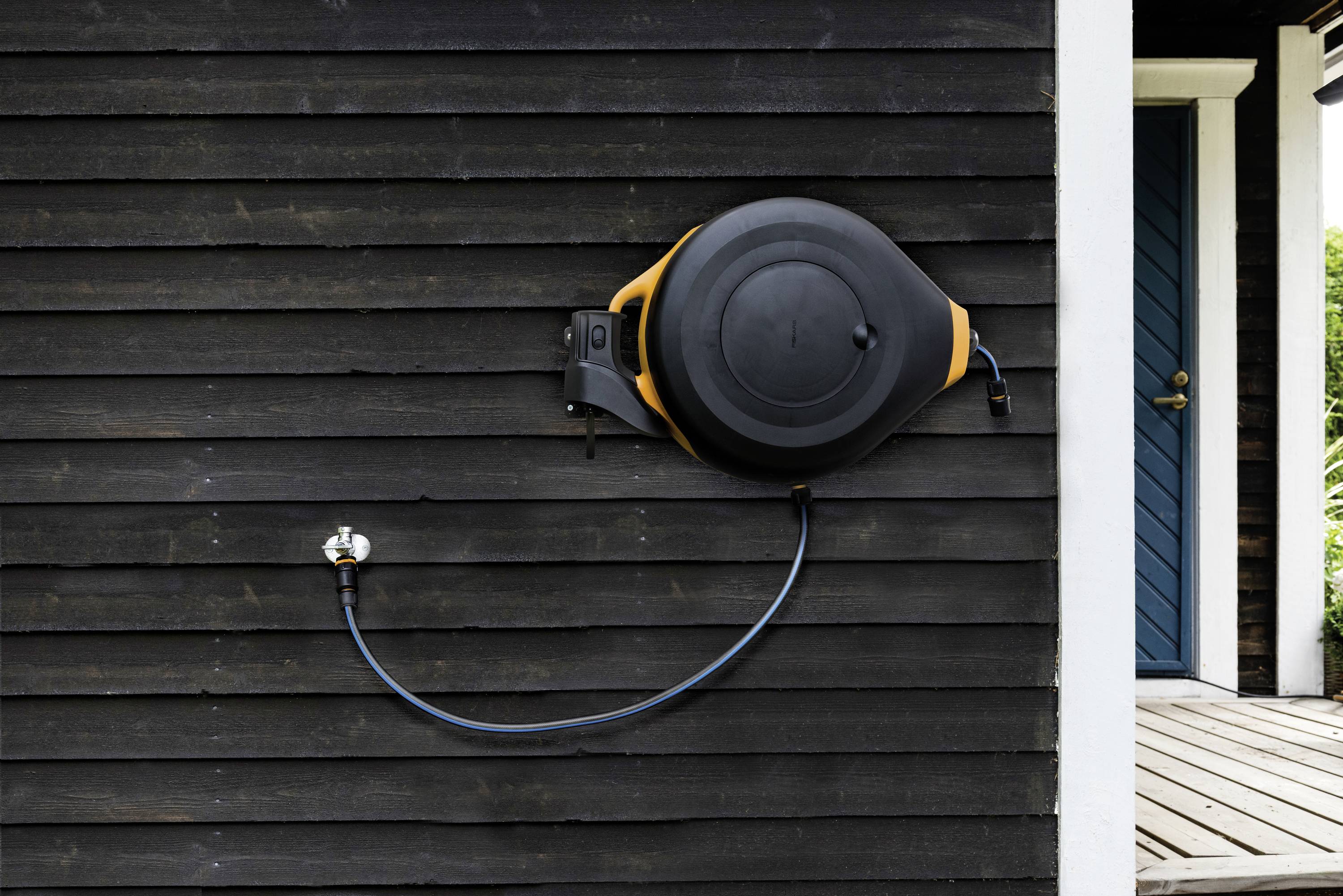Ein schwarzer Gartenschlauch ist an einer dunkel gestrichenen Holzhauswand montiert. Ein Wasserhahn ist darunter an der Wand befestigt.