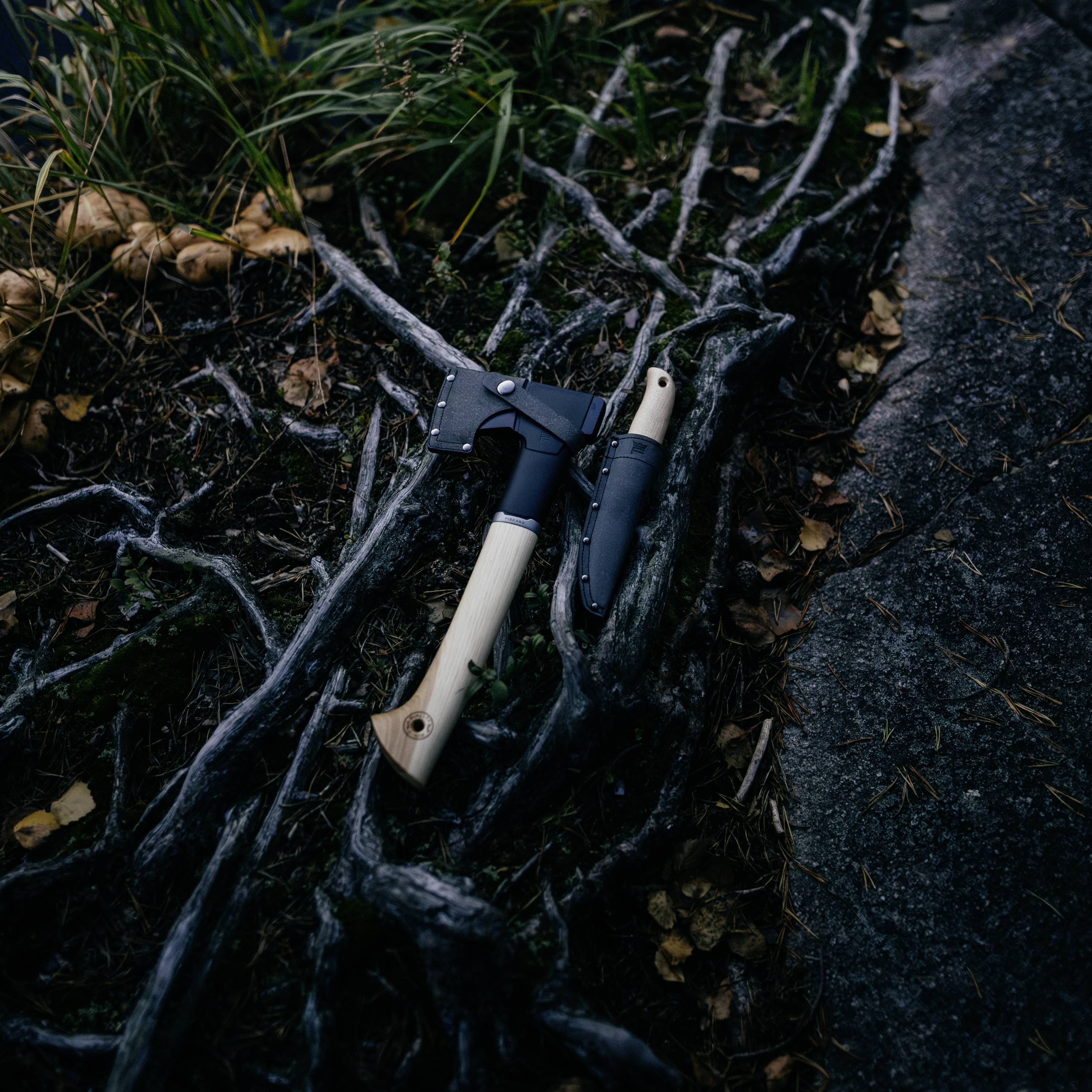 Ein Beil und ein Messer liegen auf dem Waldboden, umgeben von Wurzeln und Blättern. Sie sind parallel platziert für Outdoor-Aktivitäten.