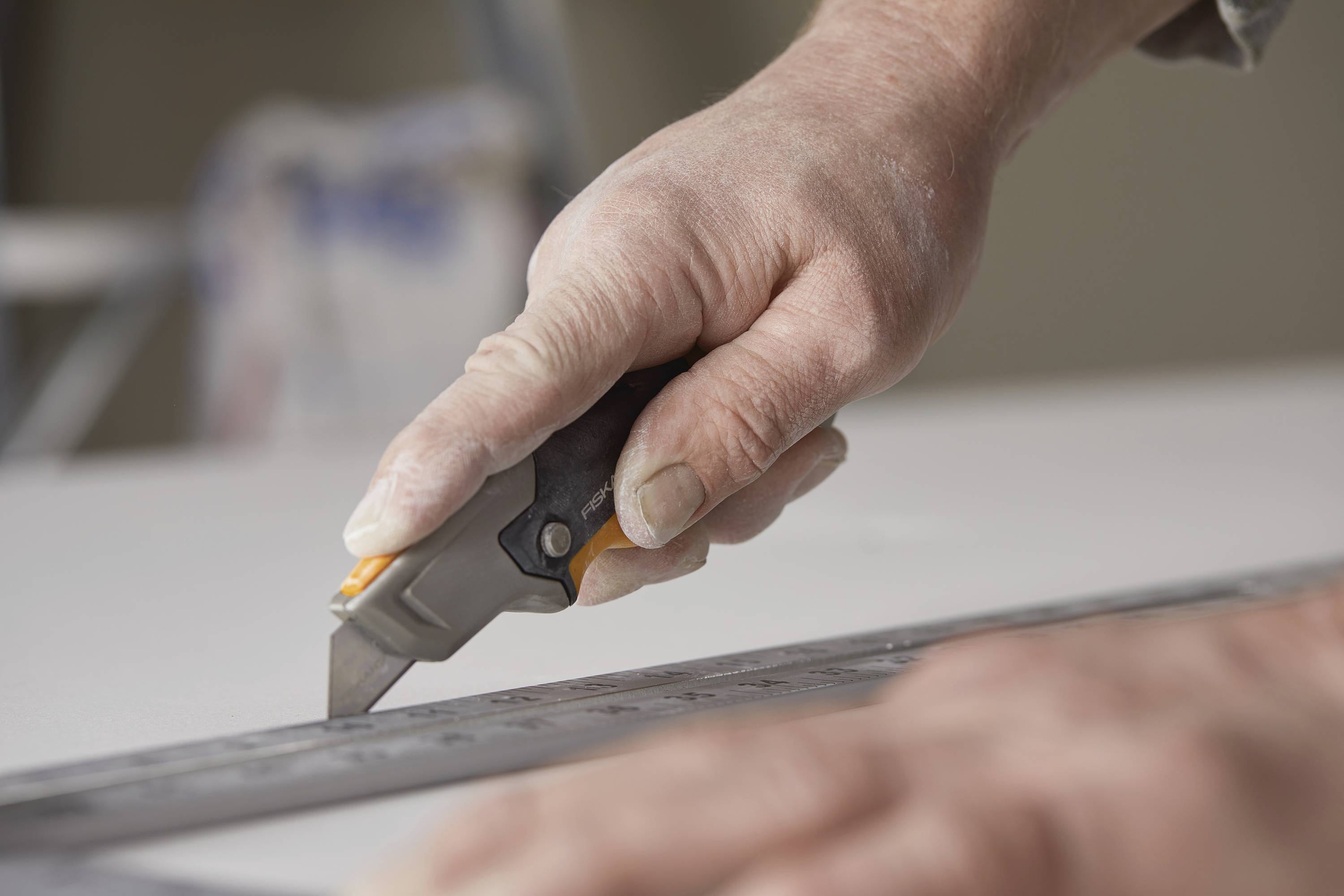 'Hand mit Cutter-Messer schneidet entlang Lineal auf weißem Untergrund.'