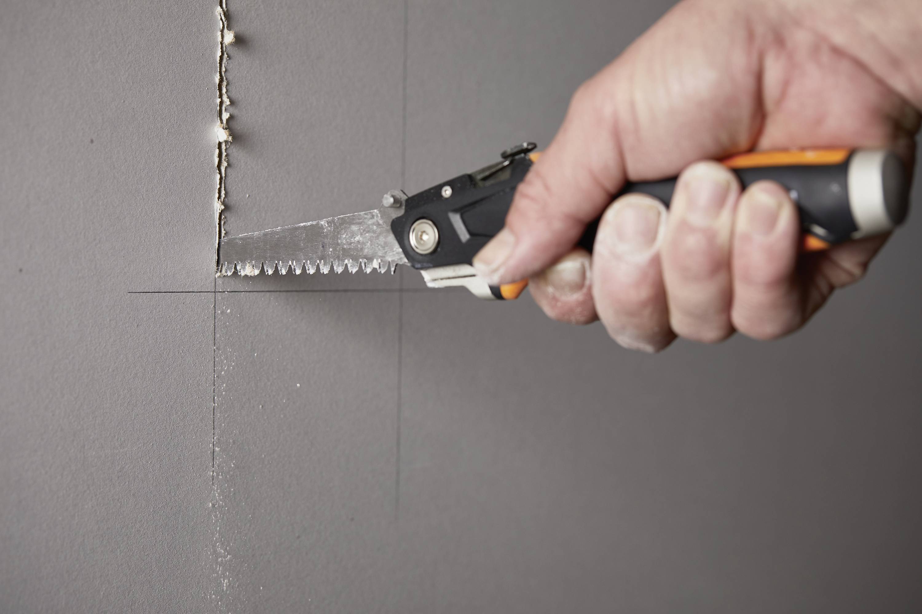 Eine Hand hält eine Säge und schneidet entlang einer Linie in eine graue Wand.