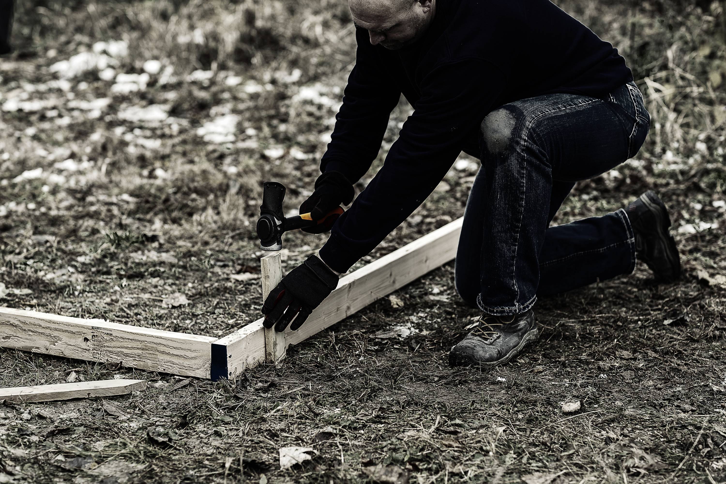Ein Mann kniet auf dem Boden und schlägt mit einem Hammer einen Holzpflock in den Boden. Er trägt dunkle Kleidung und Arbeitsschuhe.