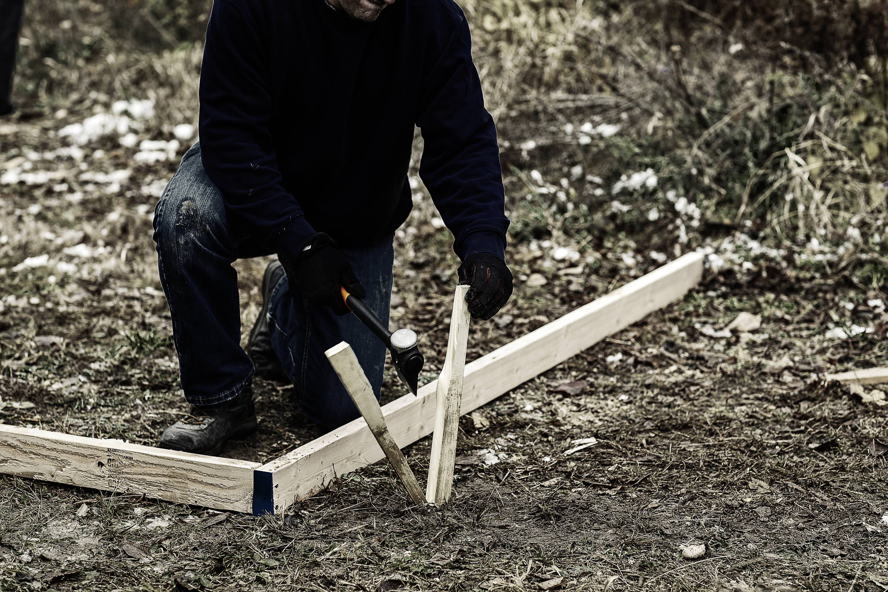 Eine Person hämmert einen Holzpfosten in den Boden neben einem Holzrahmen auf einer outdoor Baustelle.