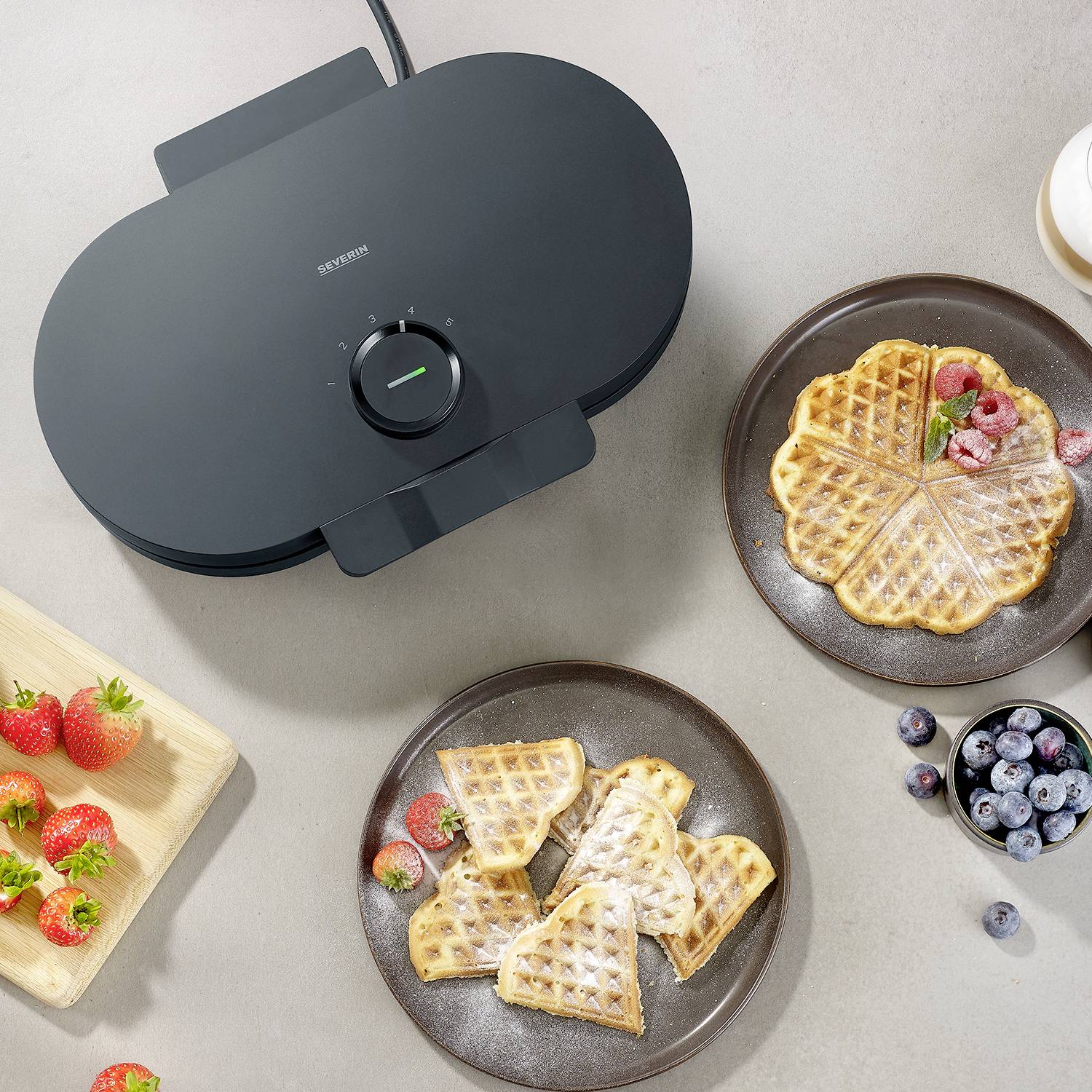Elektrisches Waffeleisen auf einem Tisch, Waffeln mit Beeren und zwei Tellern; frische Erdbeeren und Blaubeeren daneben.