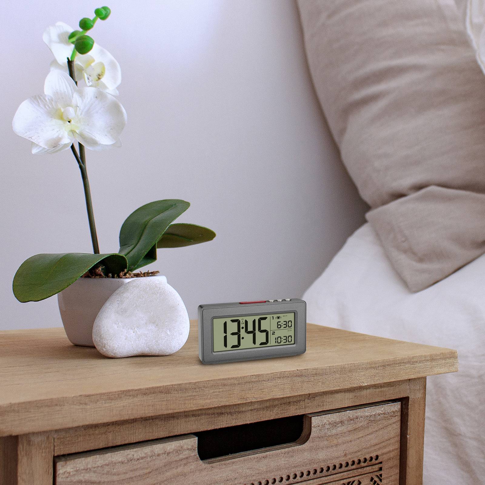 Eine weiße Orchidee in einem runden Topf steht auf einem Holznachttisch neben einem digitalen Wecker, der 13:45 Uhr anzeigt.