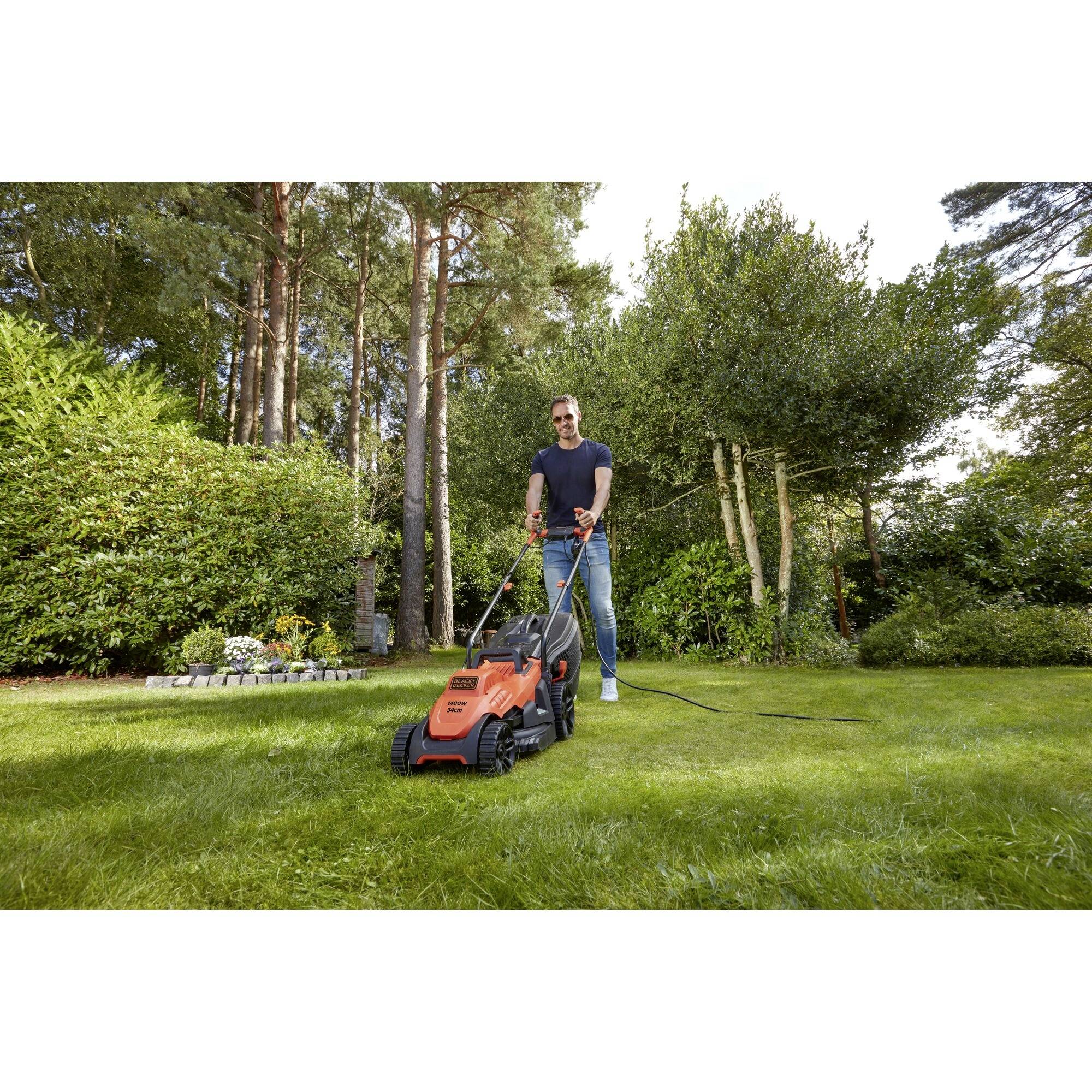 Ein Mann mäht den Rasen mit einem elektrischen Rasenmäher in einem grünen Garten, umgeben von Bäumen und Büschen.