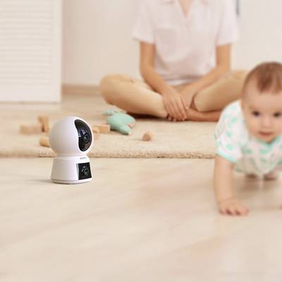 Ein Baby krabbelt auf einem Teppich, während eine Sicherheitskamera im Vordergrund fokussiert ist. Im Hintergrund sitzt eine Person.
