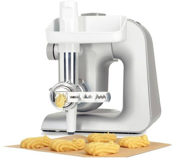 Eine silberne Küchenmaschine extrudiert Teig durch eine Spritztülle, um gezackte Kekse auf einem Backpapier zu formen.