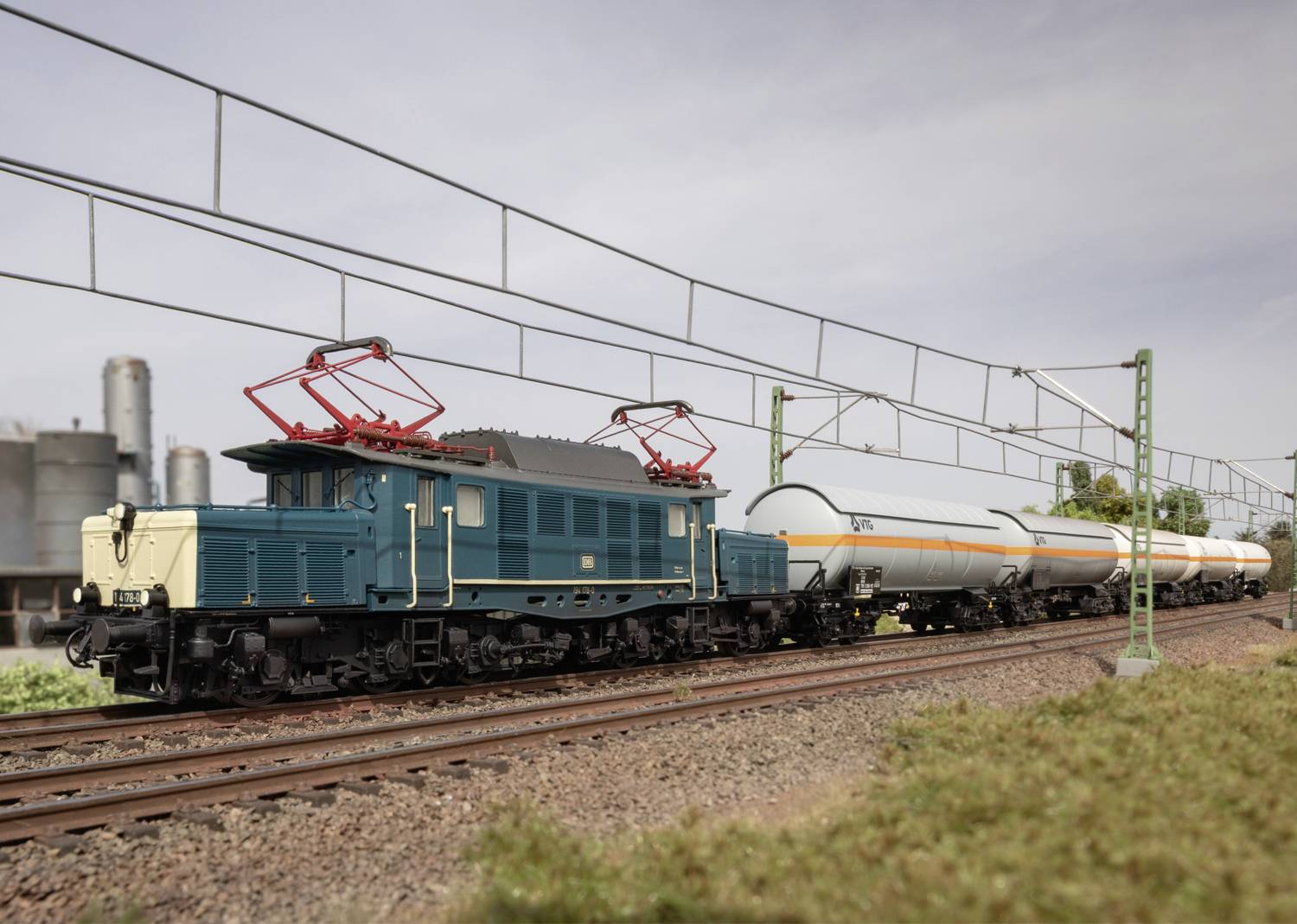 Ein Modellzug mit einer blauen Lokomotive zieht mehrere Kesselwagen auf einer Miniatureisenbahnanlage, umgeben von einer Industrieanlage.