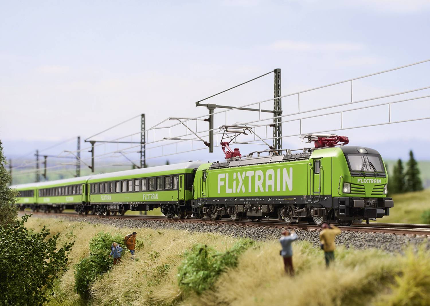 Ein grüner Zug mit der Aufschrift 'FLIXTRAIN' fährt auf einer Bahnstrecke durch eine ländliche Landschaft mit zwei Personen im Vordergrund.
