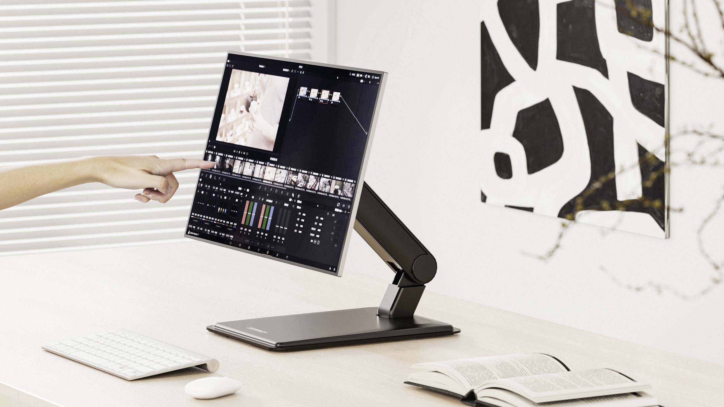 Eine Person bearbeitet ein Video auf einem Computermonitor und zeigt auf den Bildschirm, während eine Tastatur, eine Maus und ein aufgeschlagenes Buch auf dem Schreibtisch liegen.