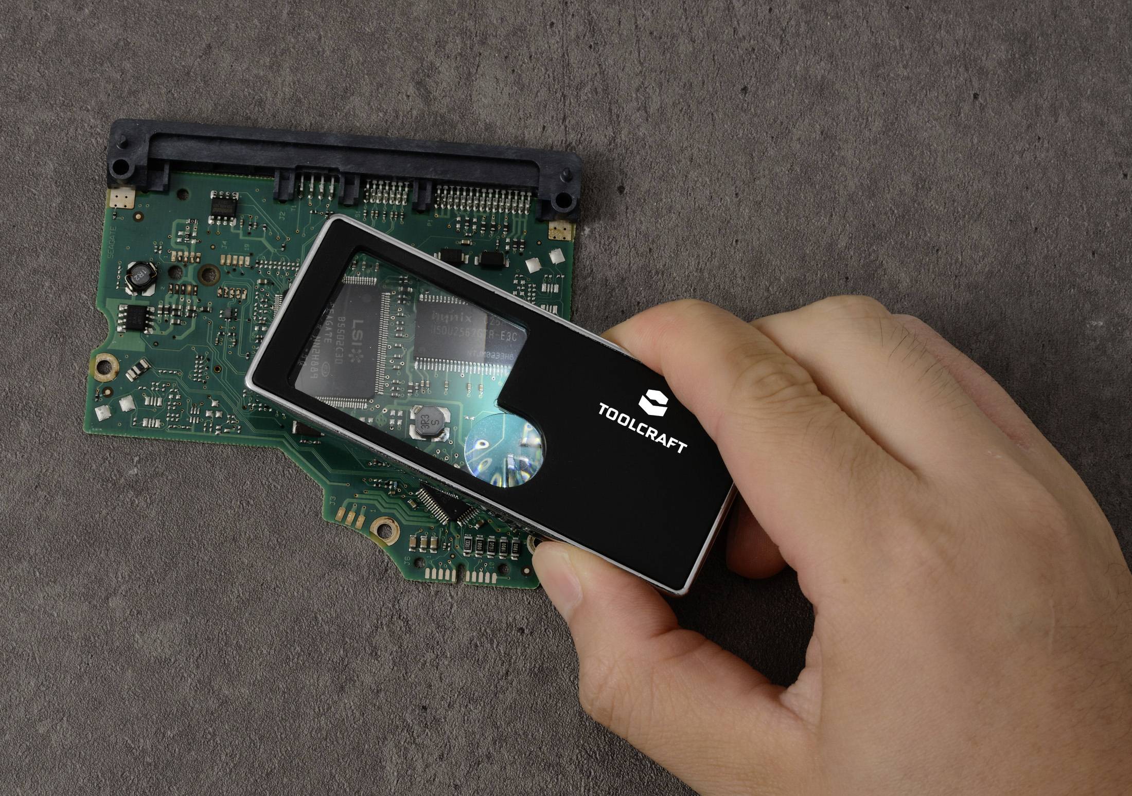 Eine Hand hält ein schwarzes Vergrößerungswerkzeug mit der Aufschrift "TOOLCRAFT" über einer grünen Leiterplatte und hebt elektronische Bauteile hervor.