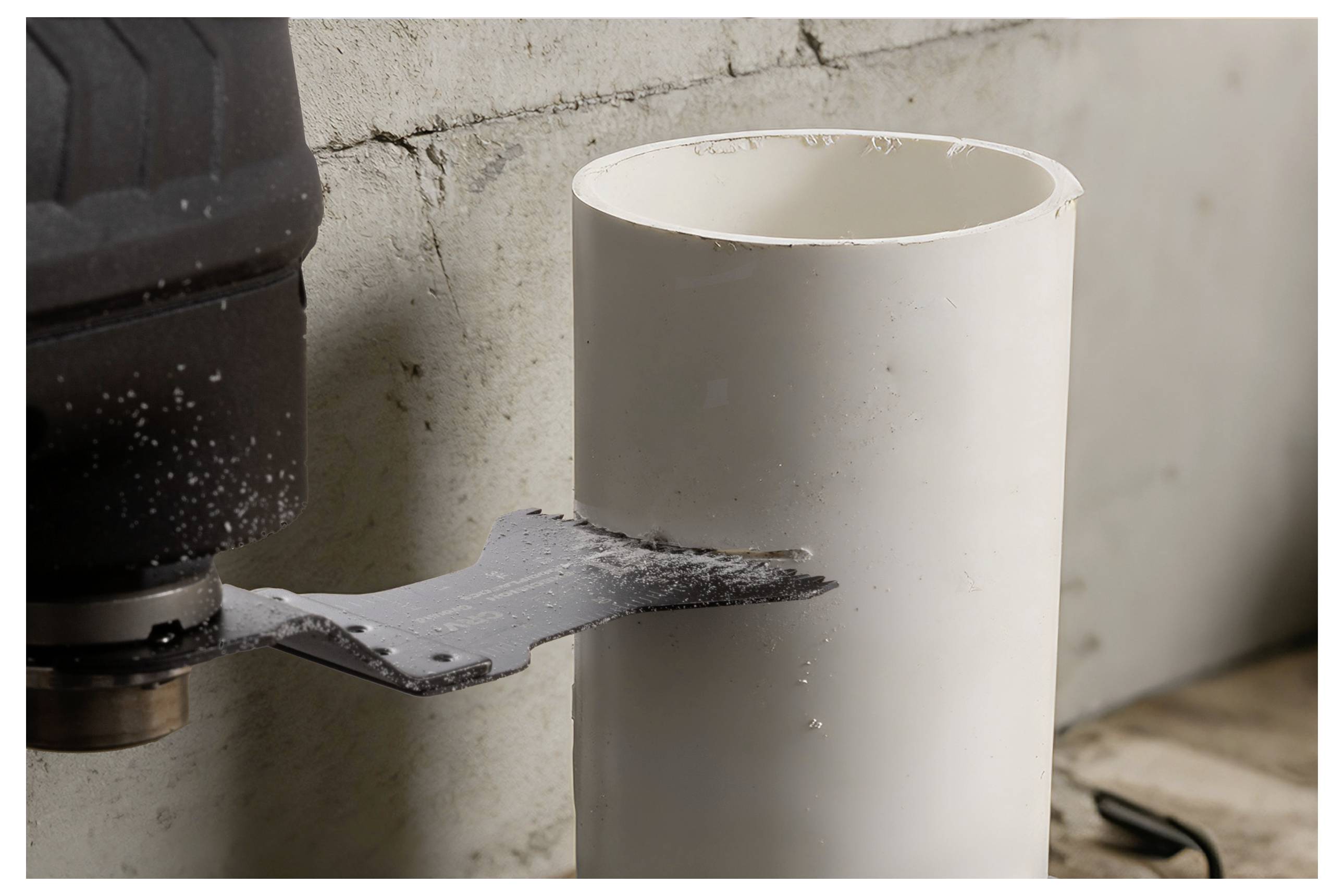 Eine Sägeblatt, das in ein vertikales weißes PVC-Rohr schneidet, mit Betonwand und Elektrowerkzeug im Hintergrund, was einen Rohrschneidevorgang veranschaulicht.