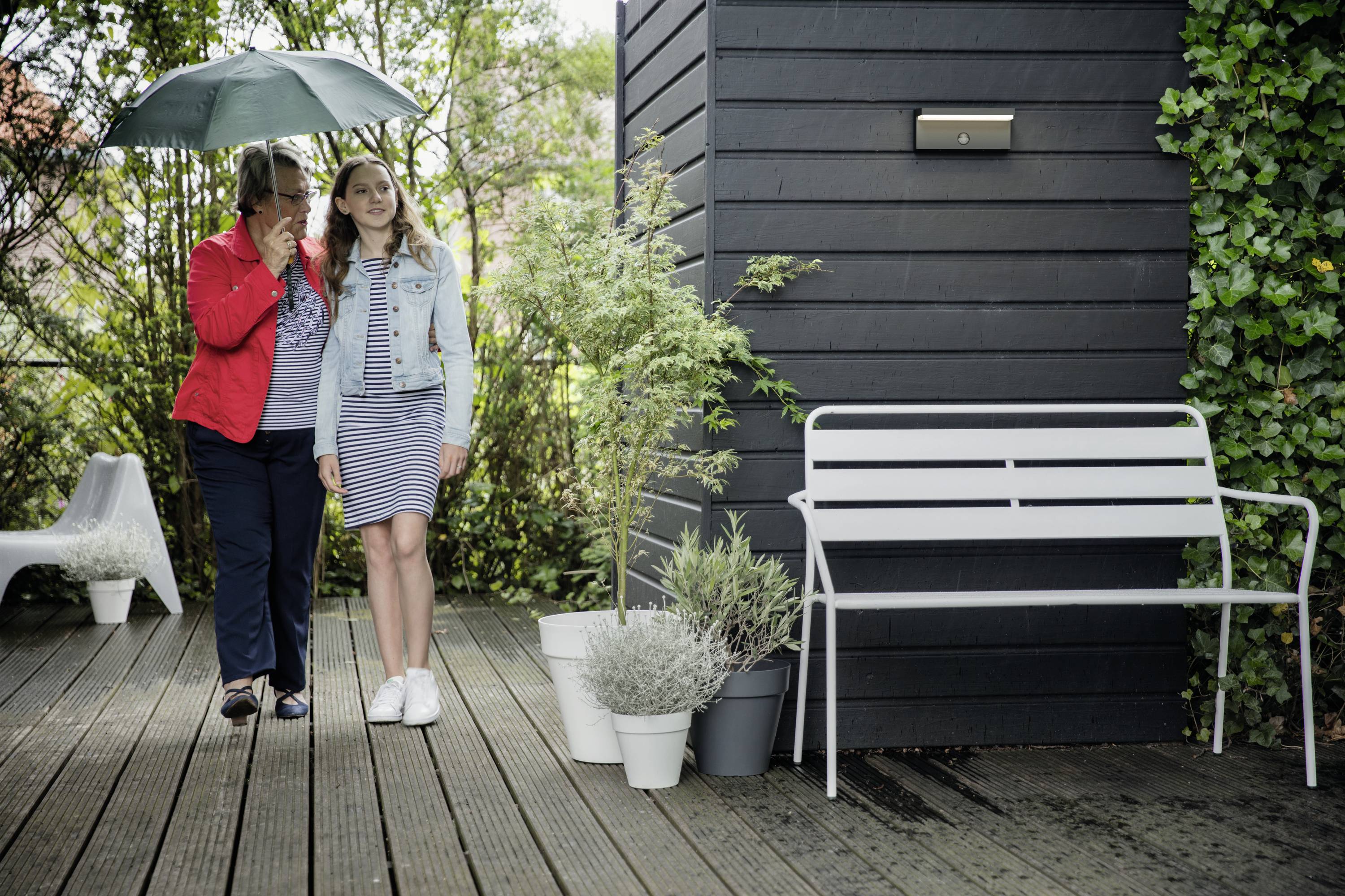 Zwei Personen unter einem Schirm stehen auf einer Holzterrasse neben einer Bank und Pflanzen. Eine Person hält den Schirm über den beiden.