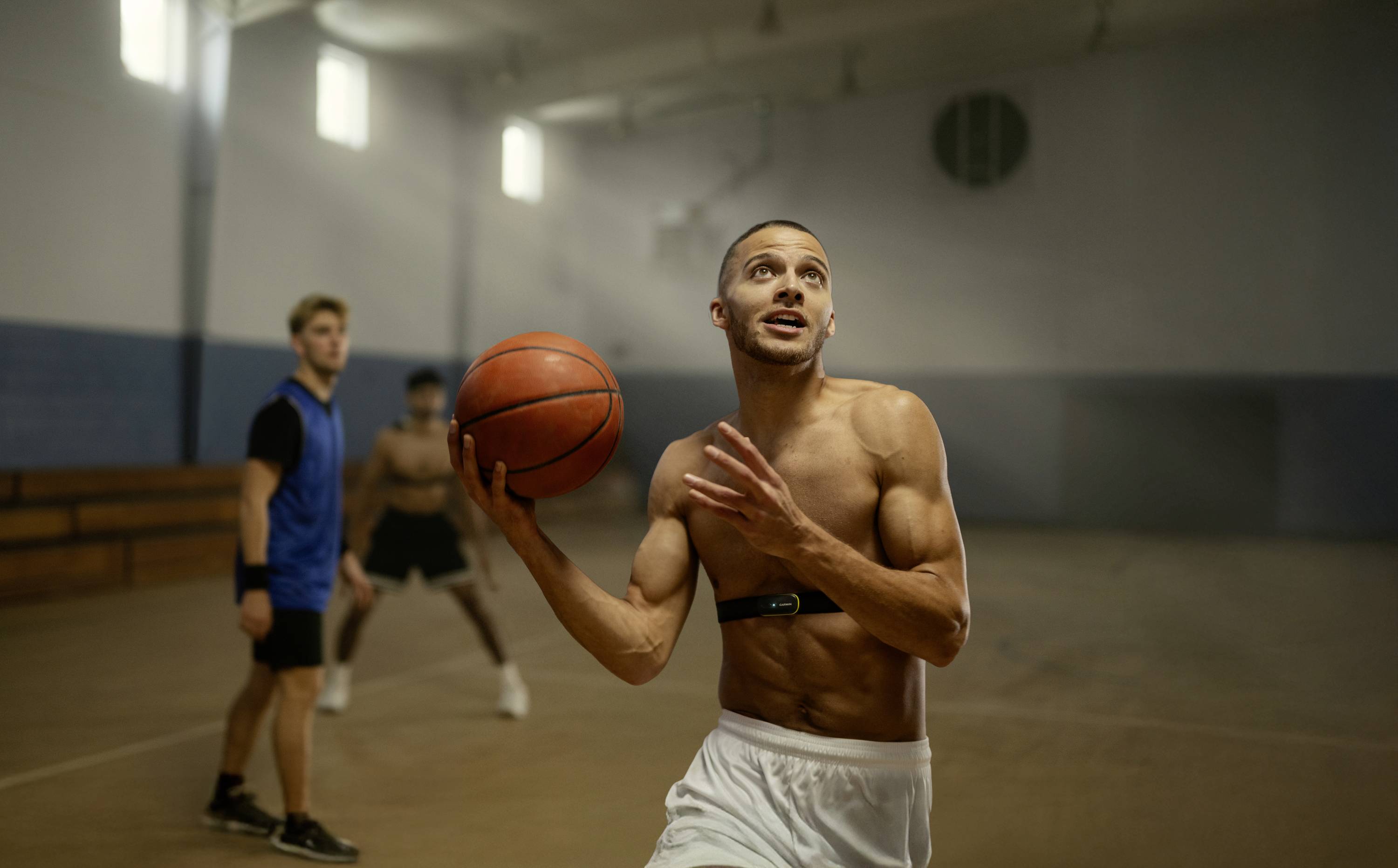Ein Mann in Sportkleidung springt mit einem Basketball in einer Turnhalle. Zwei andere Personen sind im Hintergrund zu sehen.